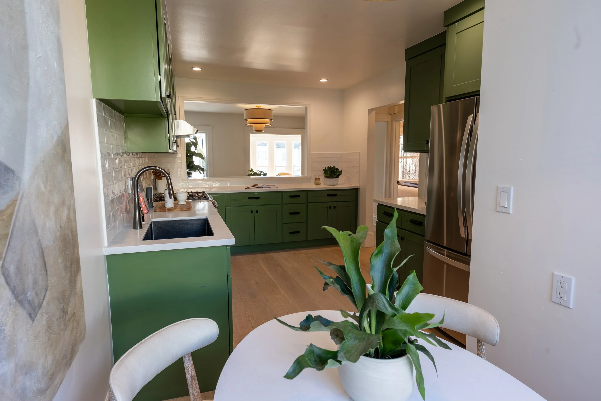 A kitchen with green cabinets, a white countertop, and a stainless steel refrigerator, seen through a window opening from a dining area with a white table and a potted plant in the foreground.