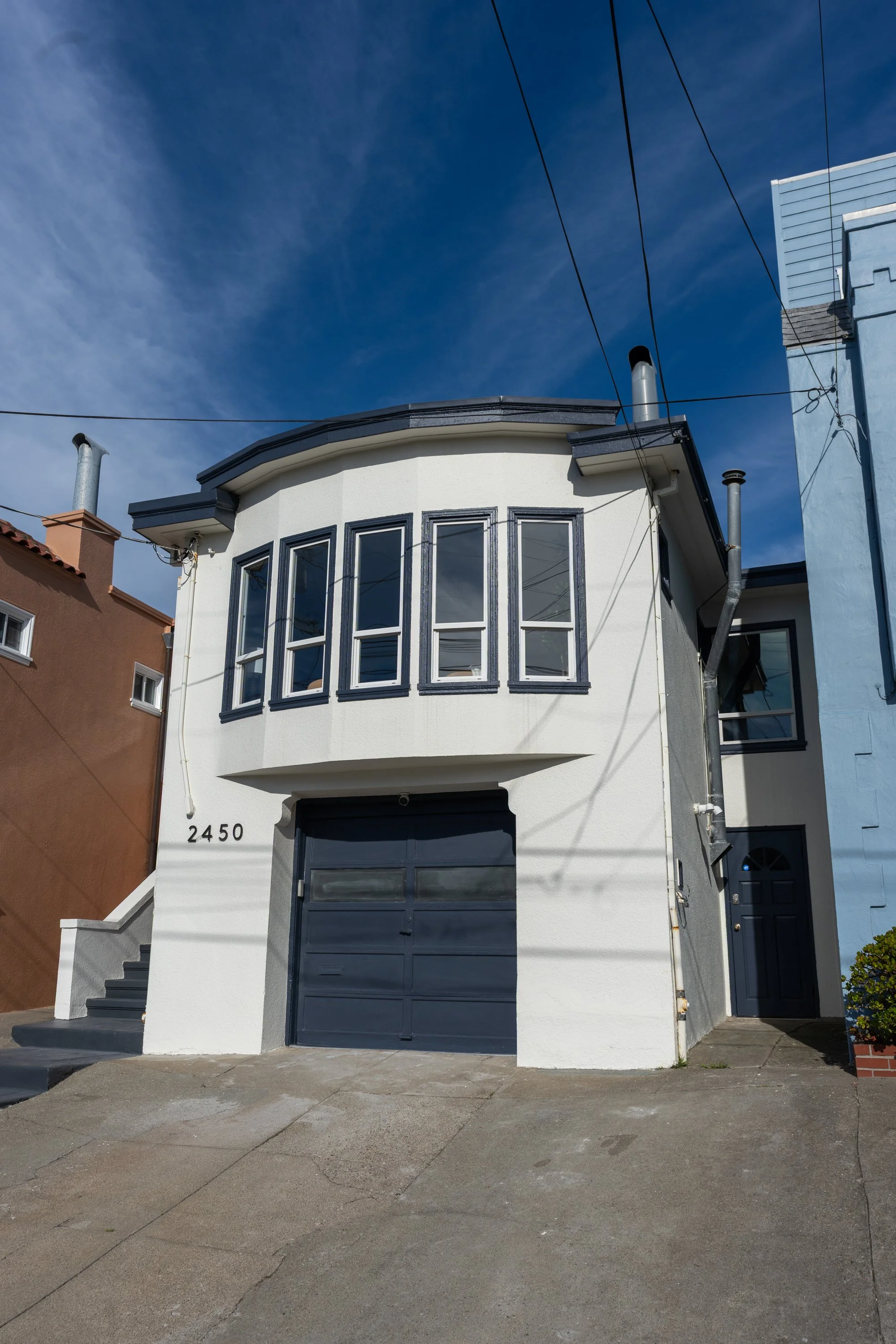 A three-story white house with black window frames and a black garage door. The house number is 2450. It features a bay window on the second floor, and there's a small staircase with a white railing leading to a black front door. The house is on a co