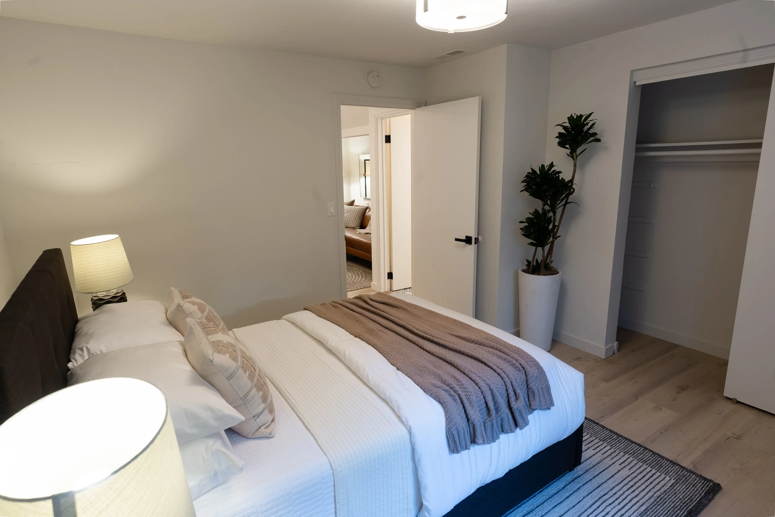 A bedroom with a neatly made bed, bedside lamps, a closet with open doors, a potted plant, and an open door leading to another room with a brown sofa and window.