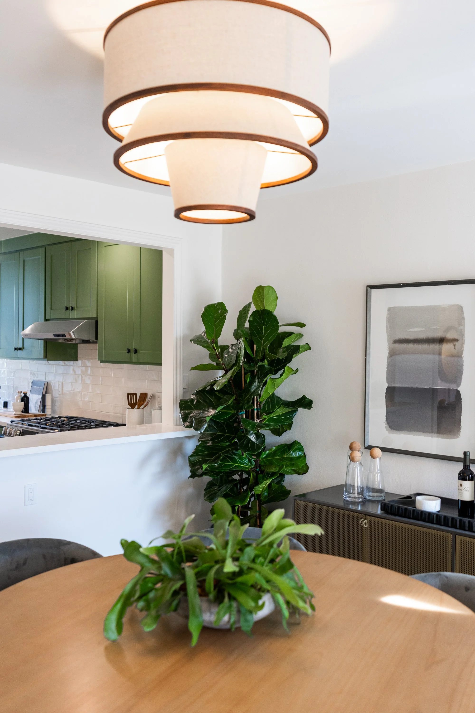 Indoor view of a modern dining and kitchen area with a hanging lamp, green cabinets, a large leafy plant, framed artwork, and a wooden table with a potted plant.