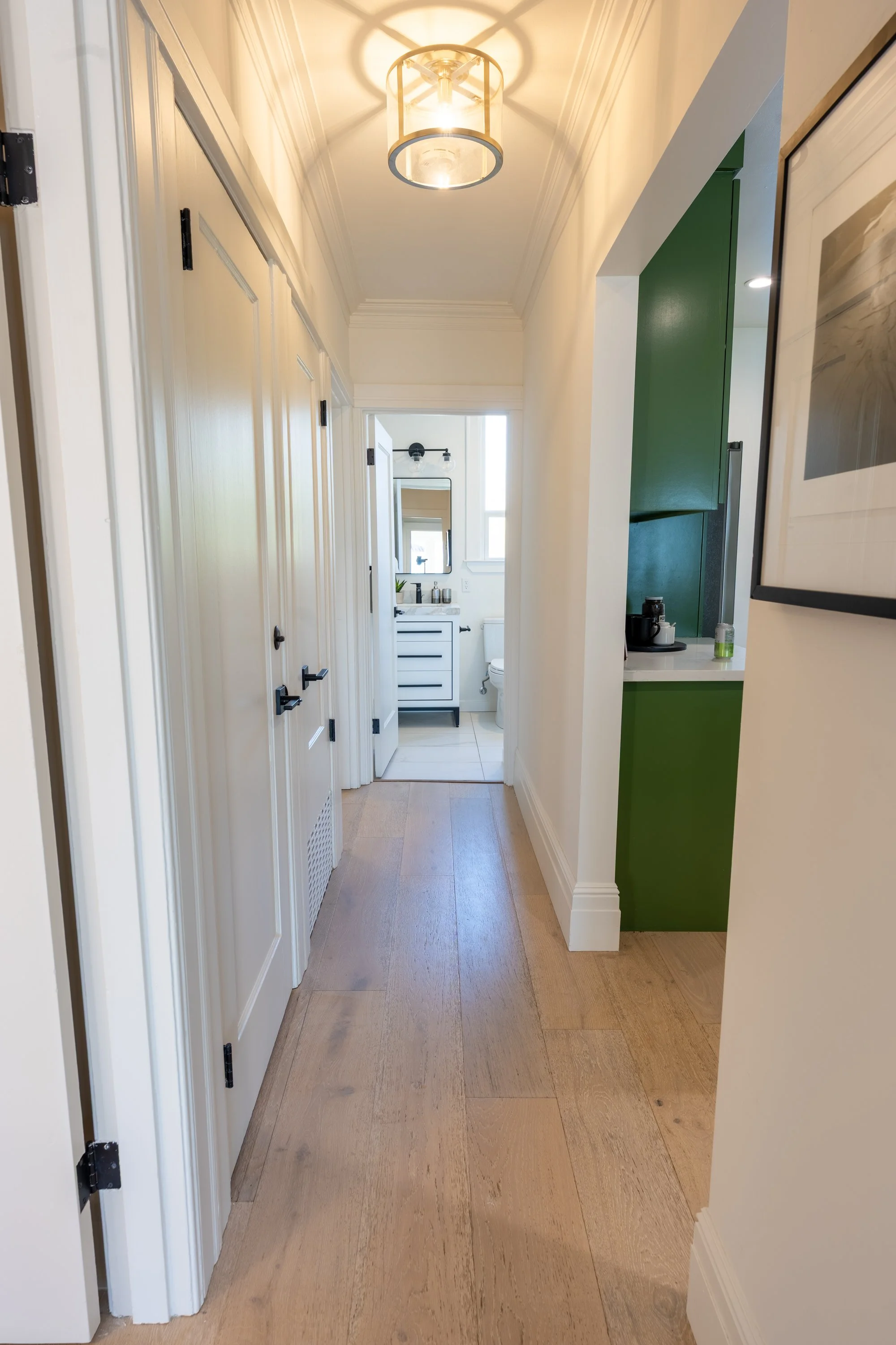 A hallway with light wood floors, white walls, and white doors on the left side, leading to a bathroom with a white vanity, black fixtures, and a mirror. A ceiling light fixture with a circular frame and globe inside illuminates the space. On the rig