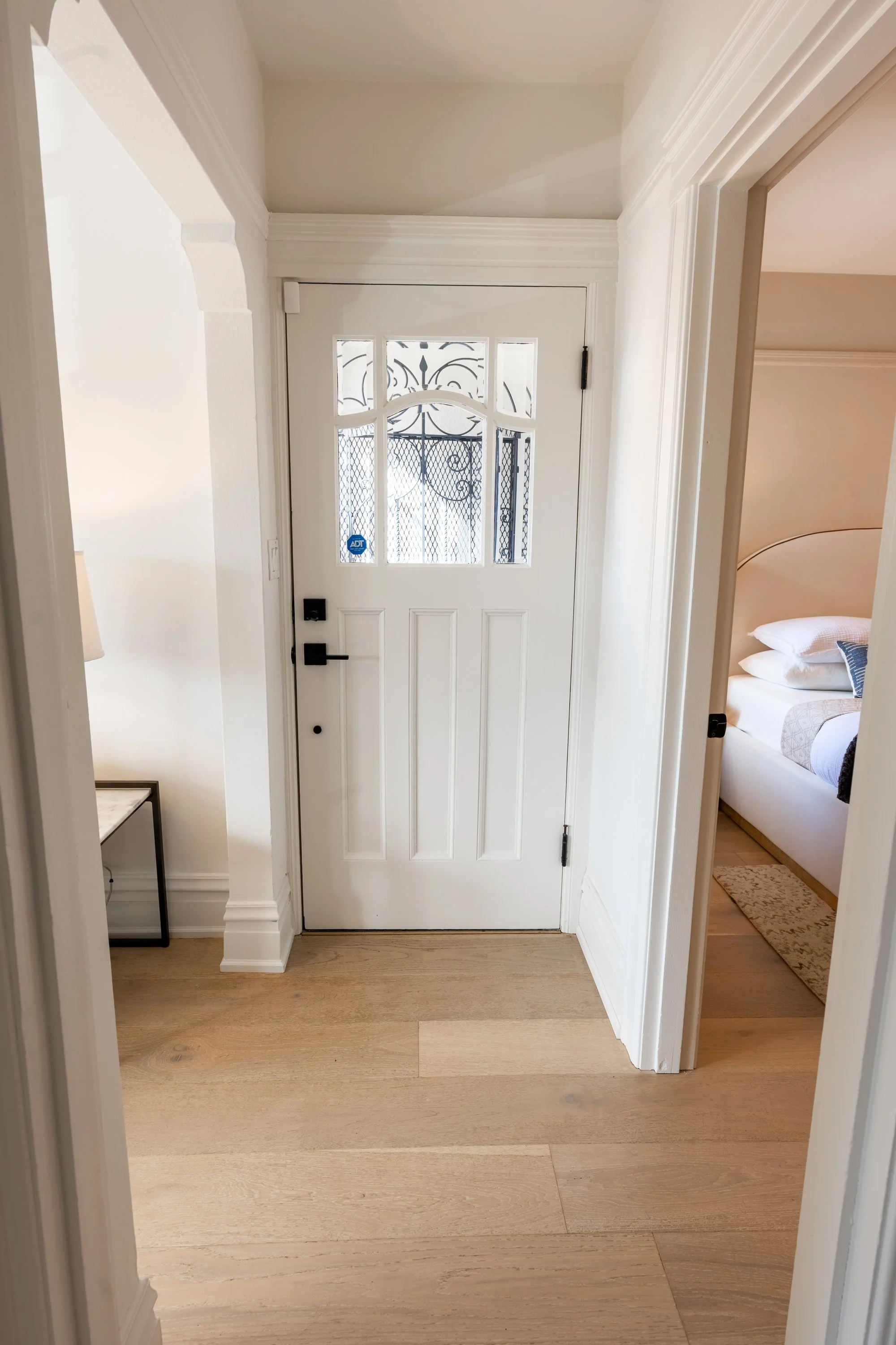 View of a white front door with glass panels and a metal security screen, seen from inside a house, with a hallway leading to a bedroom on the right.