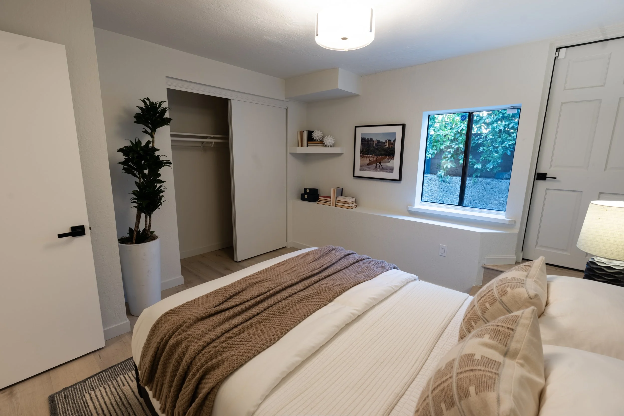 A cozy bedroom with a bed covered in white linens and a brown throw blanket. There is a window with a view of outdoor greenery and a closet door slightly open. Decorative items like framed pictures, books, and white star-shaped ornaments are on a she