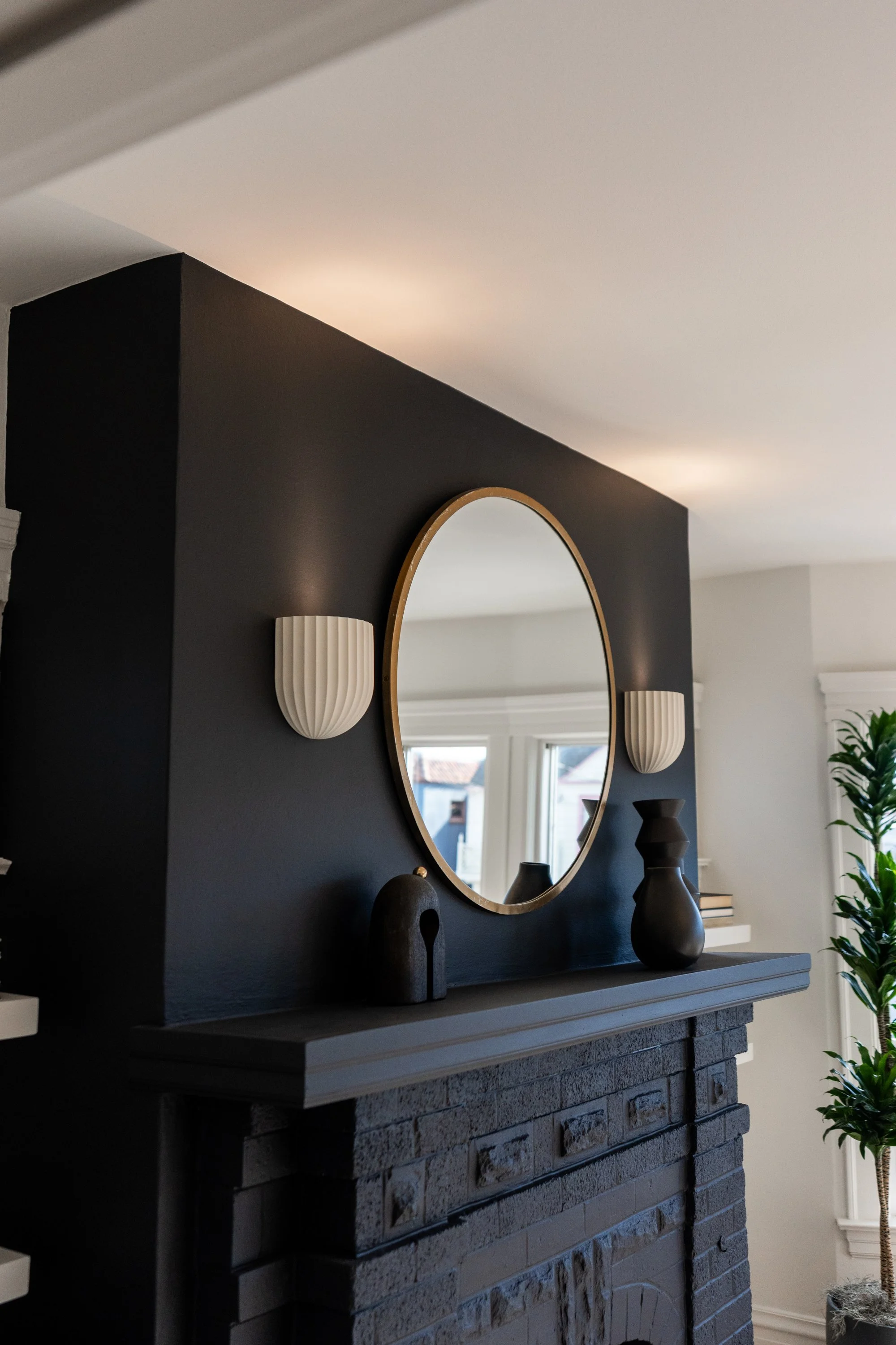 Living room fireplace with black painted wall, round mirror, white wall sconces, and decorative vases on the mantel. A window and plant are visible in the background.