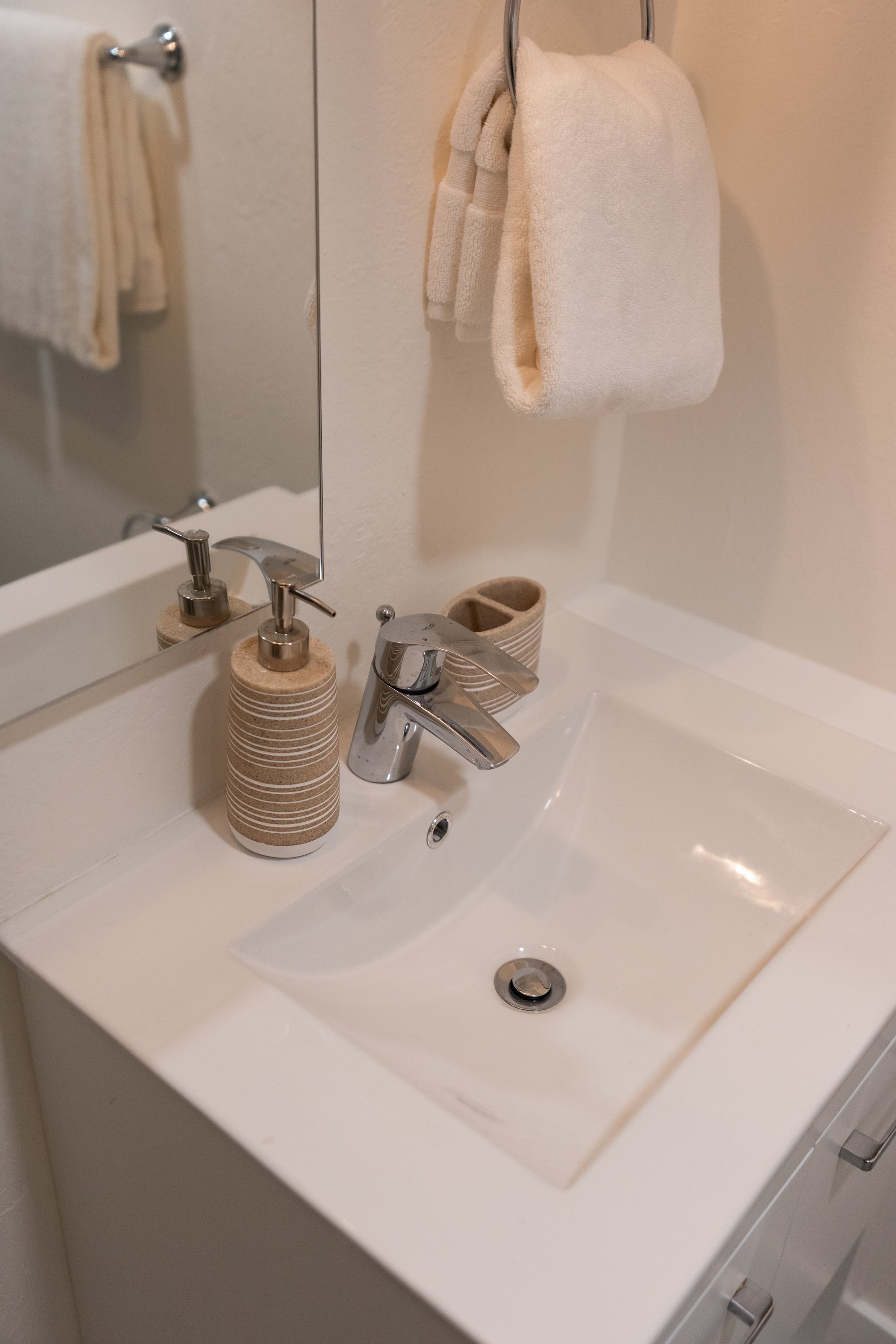 A bathroom sink with a soap dispenser, a toothbrush holder, and a mirror. Towels hang on the wall, one on a ring and another on a bar.