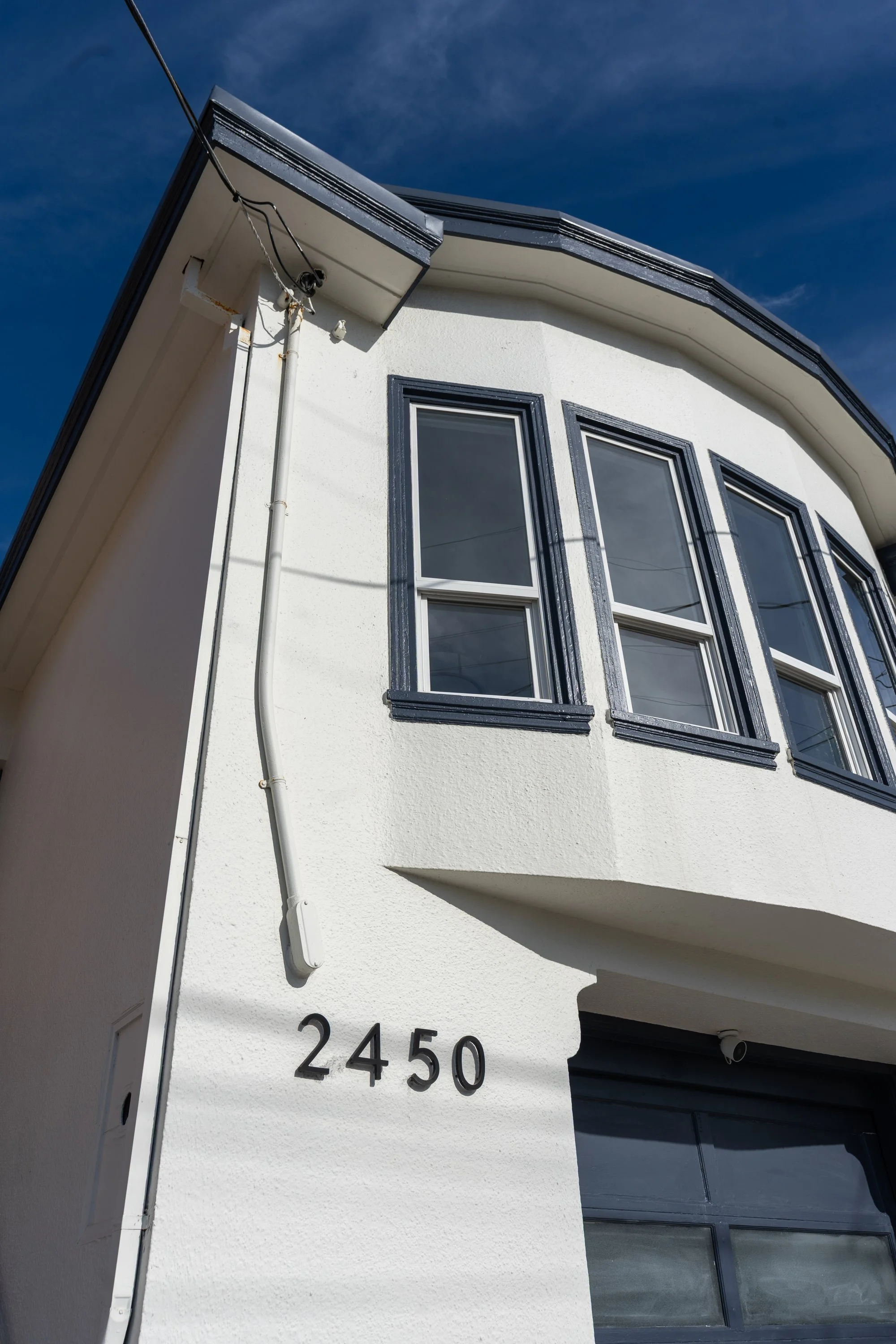 Close-up of the corner of a white house showing windows with black trim, a gutter system, and house number 2450 on the wall, under a blue sky.