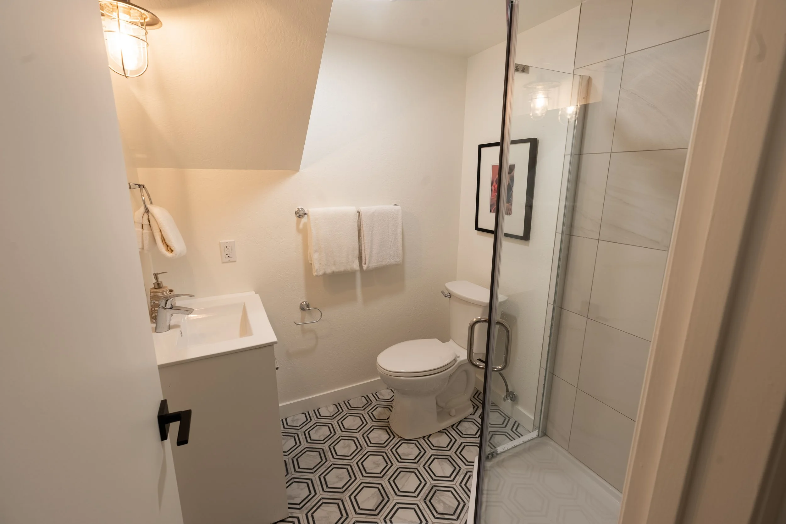 A small bathroom with white walls, a toilet, a small white sink with a simple faucet, and a towel rack with two towels. There is a framed picture on the wall, a lamp with a cage-style cover, and a glass shower enclosure with white tiles inside. The f