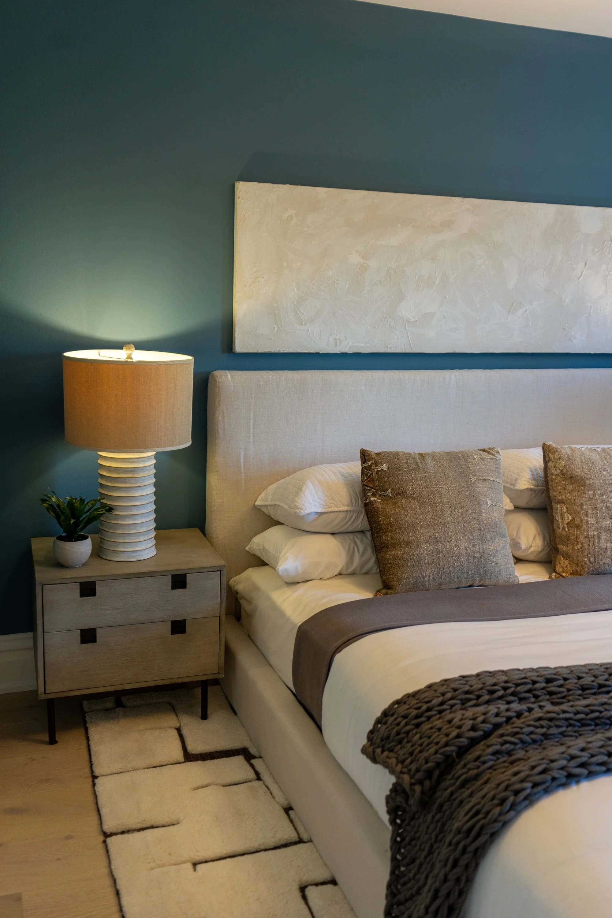 A bedroom with a bed, pillows, a beige headboard, a nightstand with a lamp and a small plant, a beige and brown rug, and dark-colored walls with a large white abstract art piece hanging above the bed.