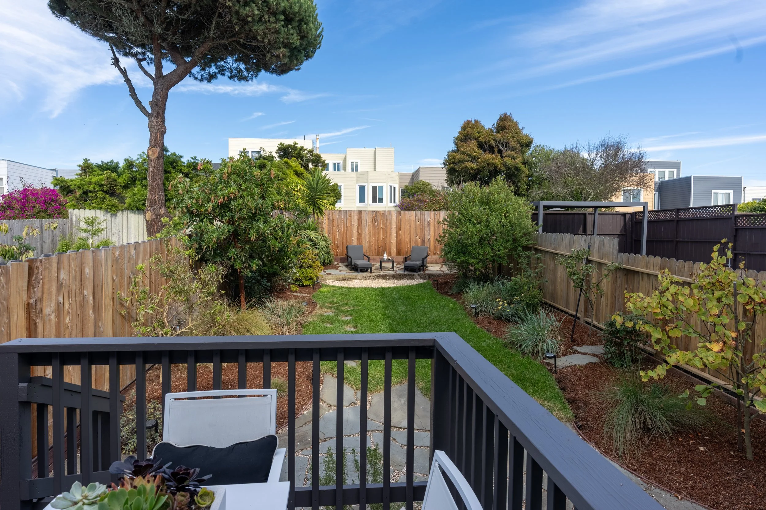 View of a backyard with a wooden fence, lush green plants, trees, two lounge chairs with a small table in between, a manicured lawn, and neighboring modern houses under a blue sky with some clouds.