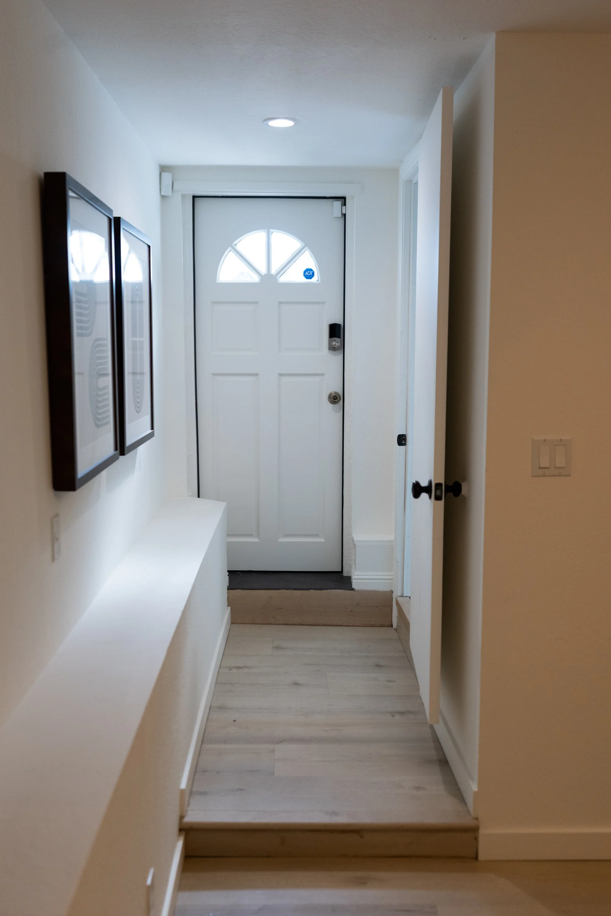 A hallway leading to a closed white door with a small semi-circular window. There are two framed pictures on the left wall and a white shelf below them. To the right, there is a doorway leading to another room. The floor has light-colored wood planks