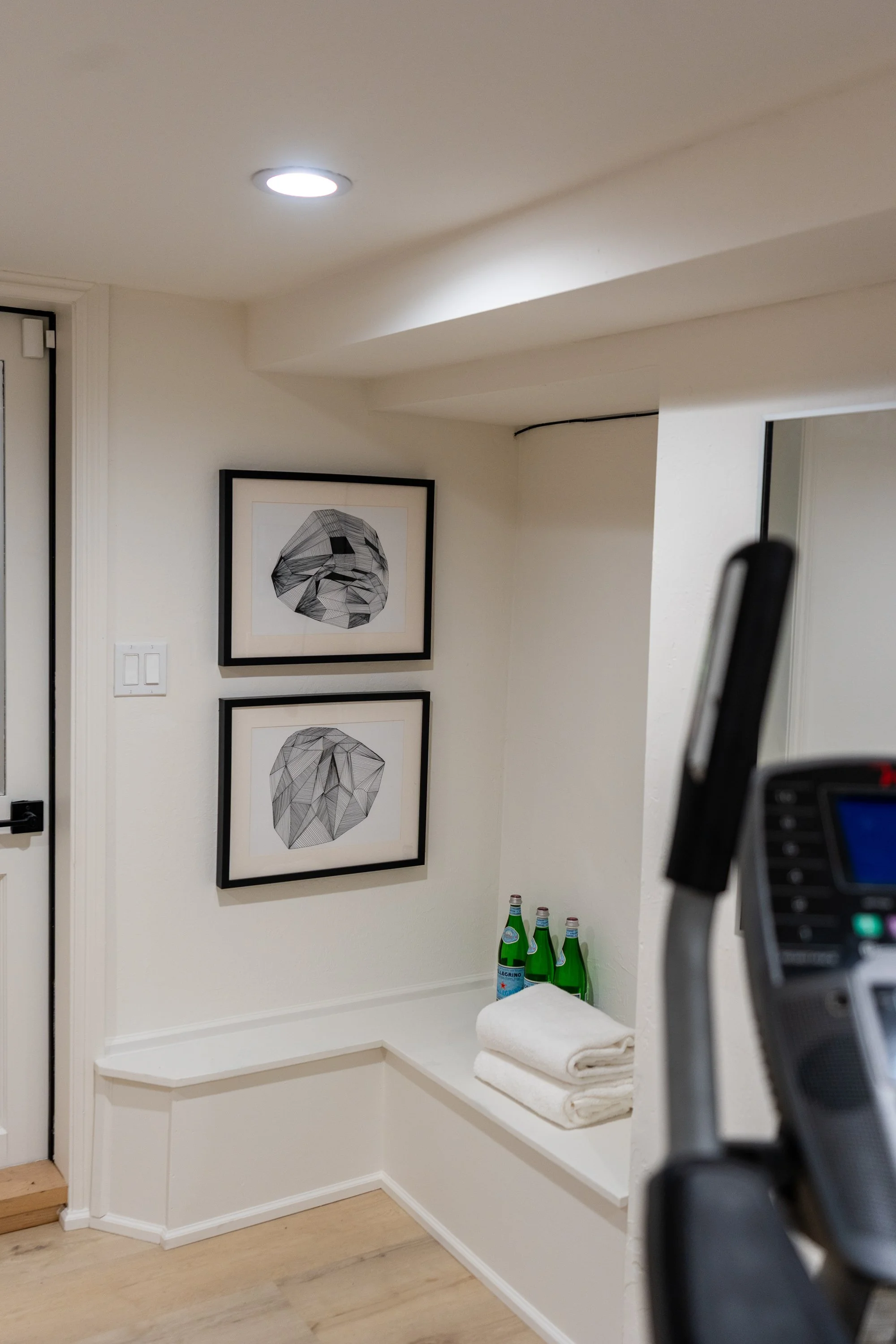 Interior white wall corner with framed abstract geometric black and white artwork, towels and bottles on a ledge, and a door frame on the left.