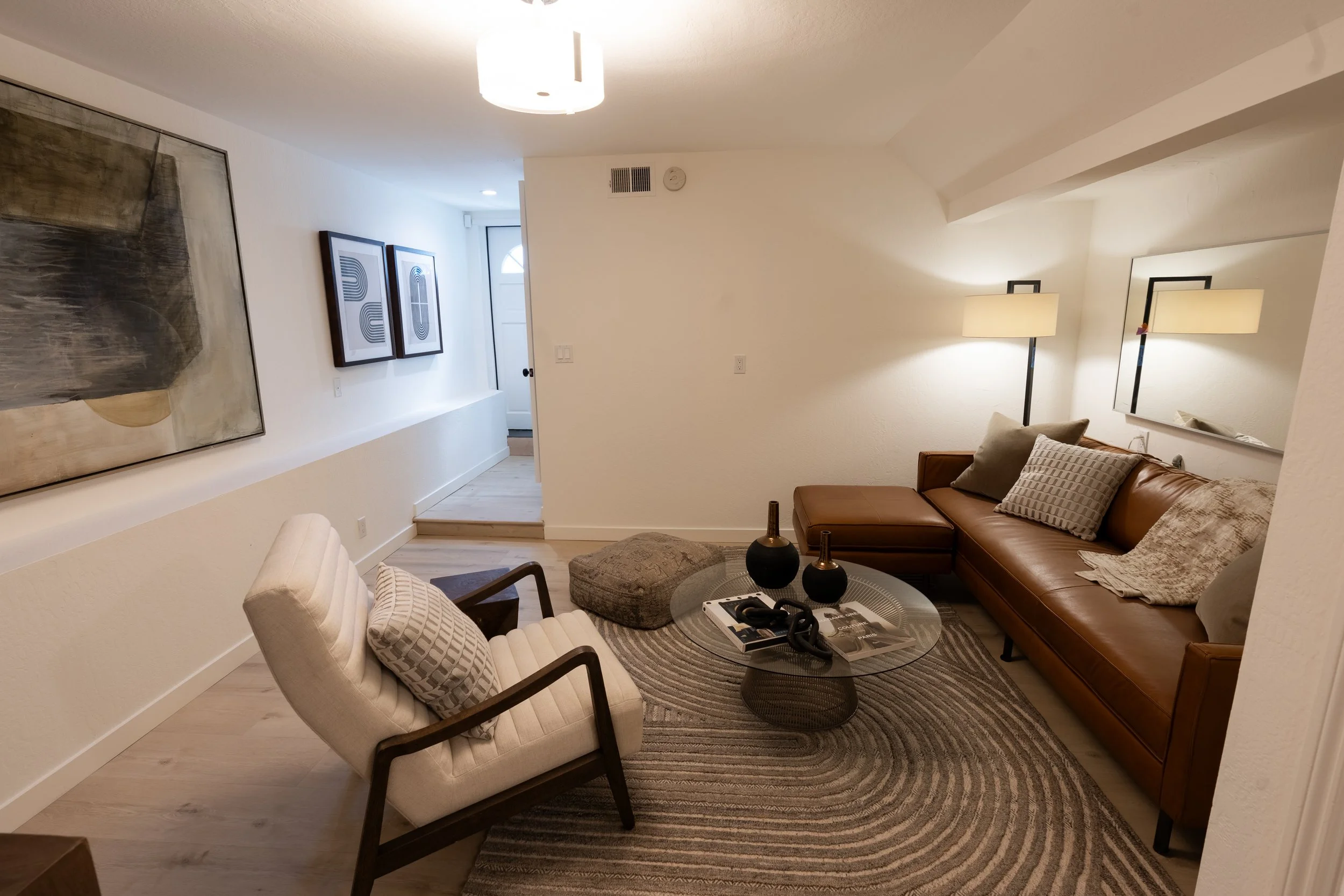 Living room with brown leather sofa, white armchair with pillows, round glass coffee table with decorative vases, wall art, and floor lamps, with white walls and light wood flooring.