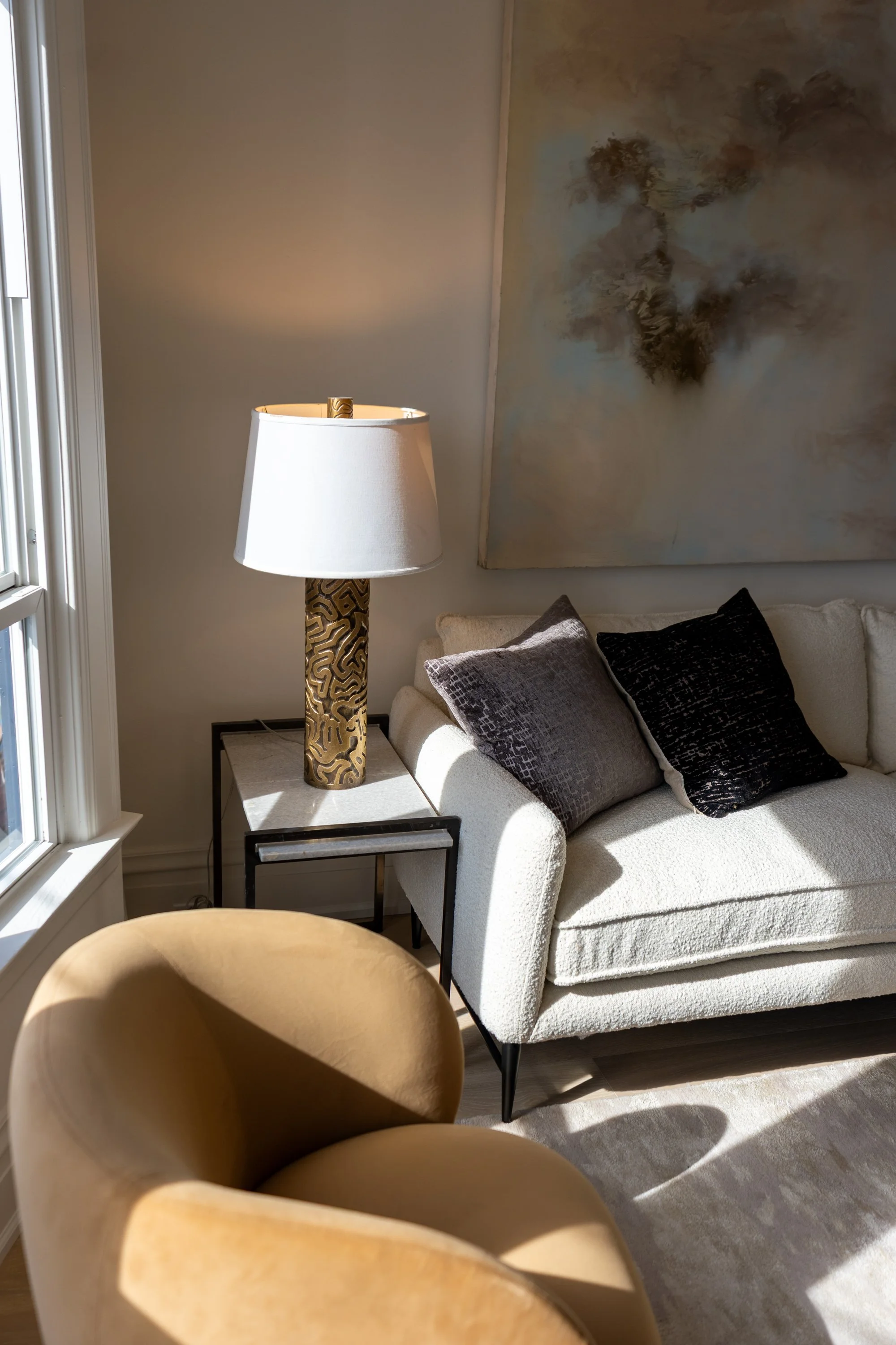 Living room corner with an off-white textured sofa, black metal side table, decorative pillows, a large abstract wall art, a unique floor lamp with a carved gold metal base and white shade, and a window with sunlight illuminating the space.