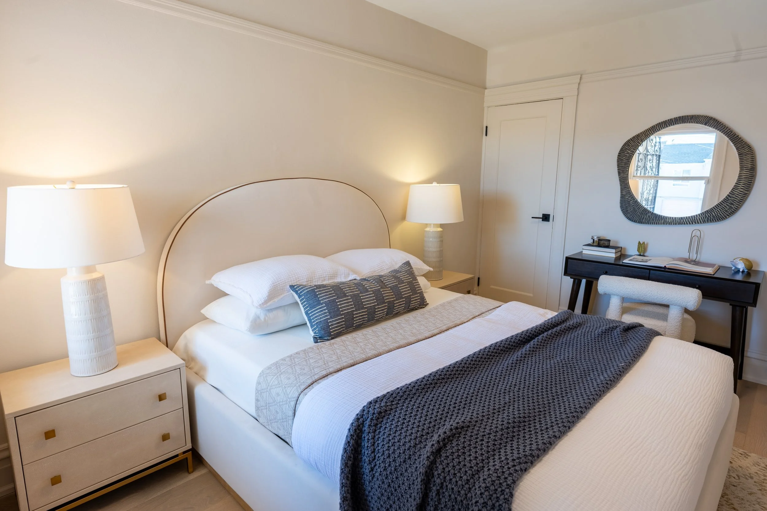 A bedroom with a bed, white and beige bedding, and a blue pillow. Two matching lamps are on nightstands on either side of the bed. A round mirror with a textured black frame hangs above a dark desk, which has a few decorative items, books, and an ope