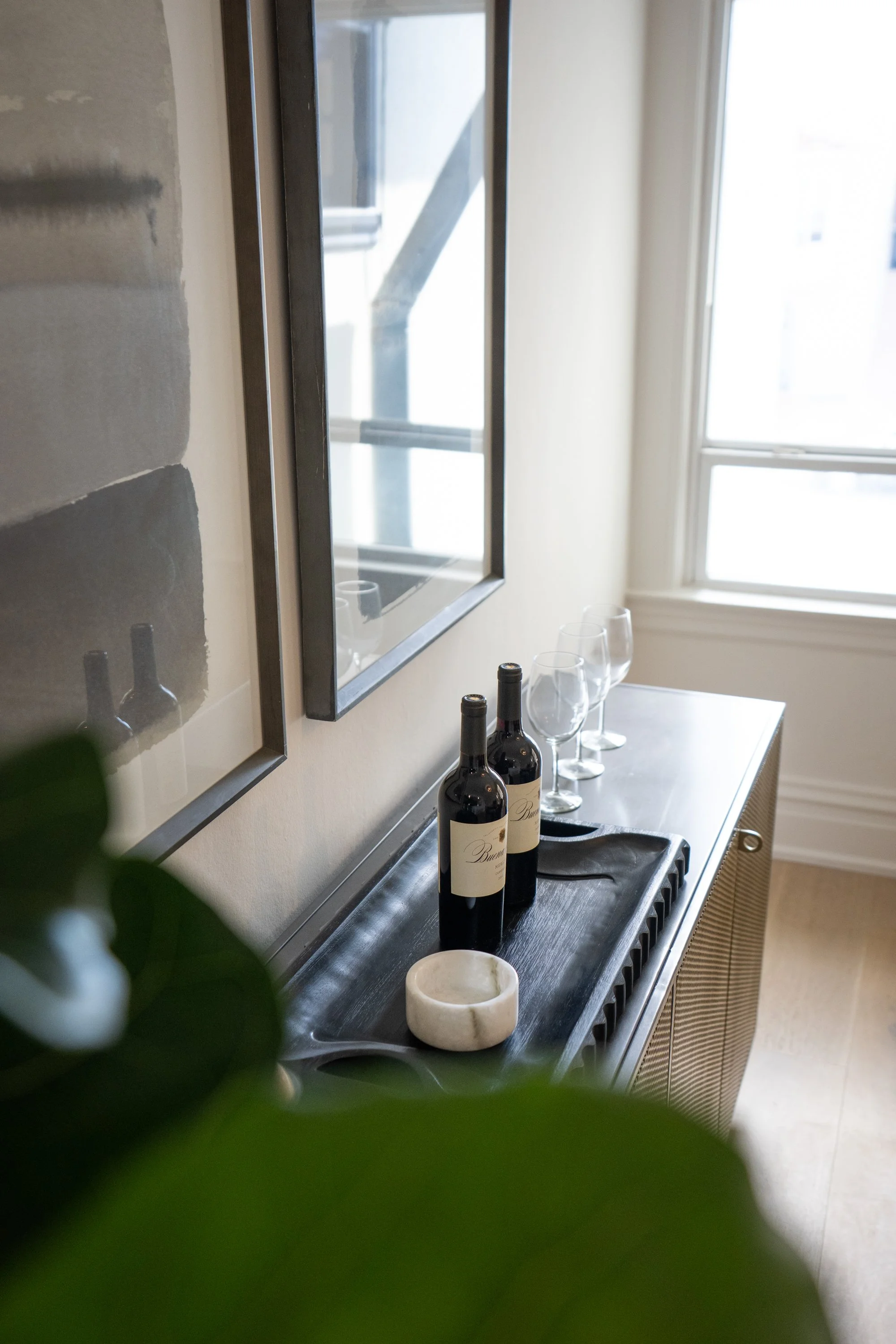 A kitchen sideboard with two wine bottles, three wine glasses, a cloth, and a marble dish, near a window with natural light.
