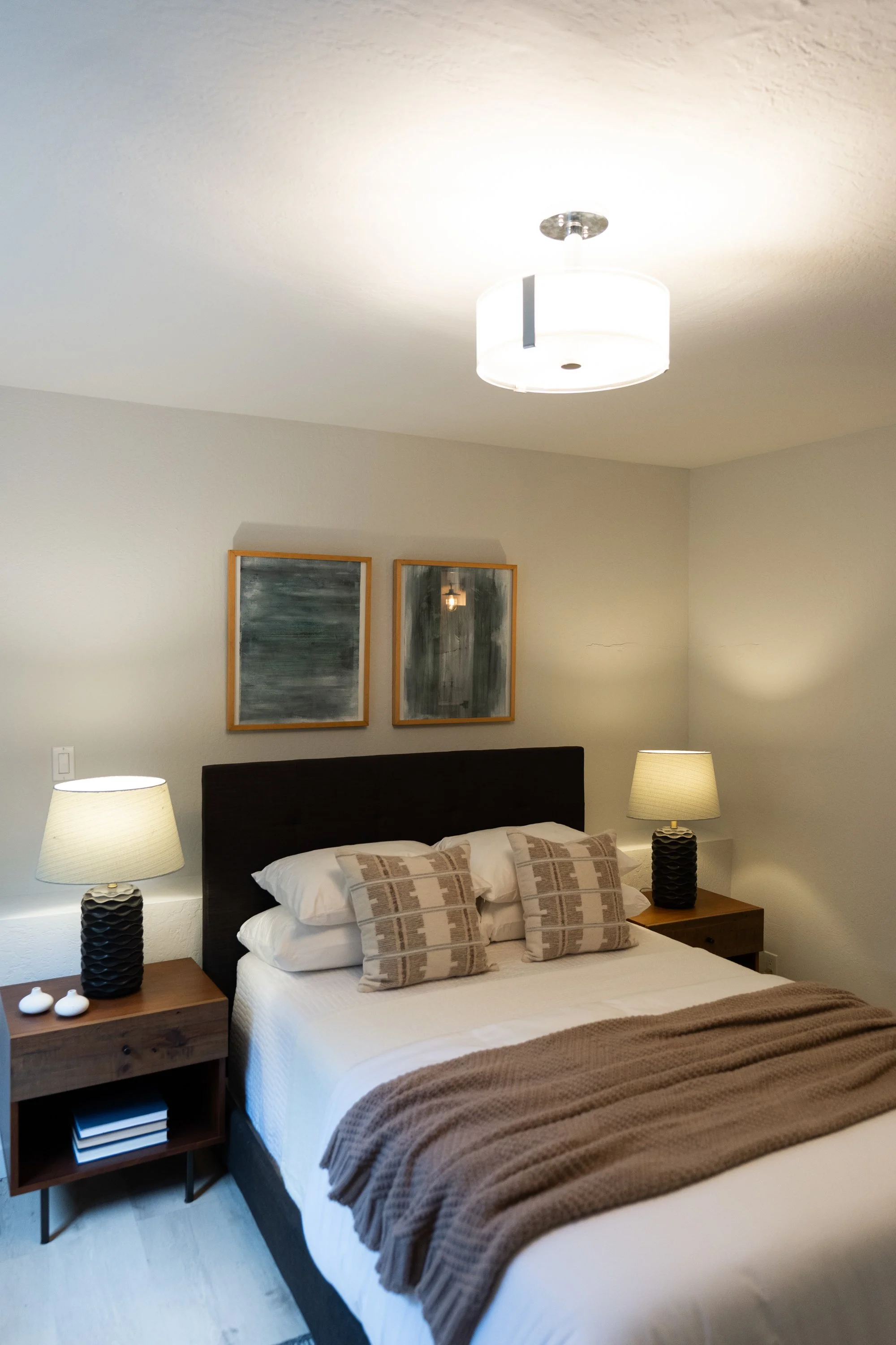 A bedroom with a bed, two lamps, two framed abstract paintings on the wall, and a ceiling light fixture.