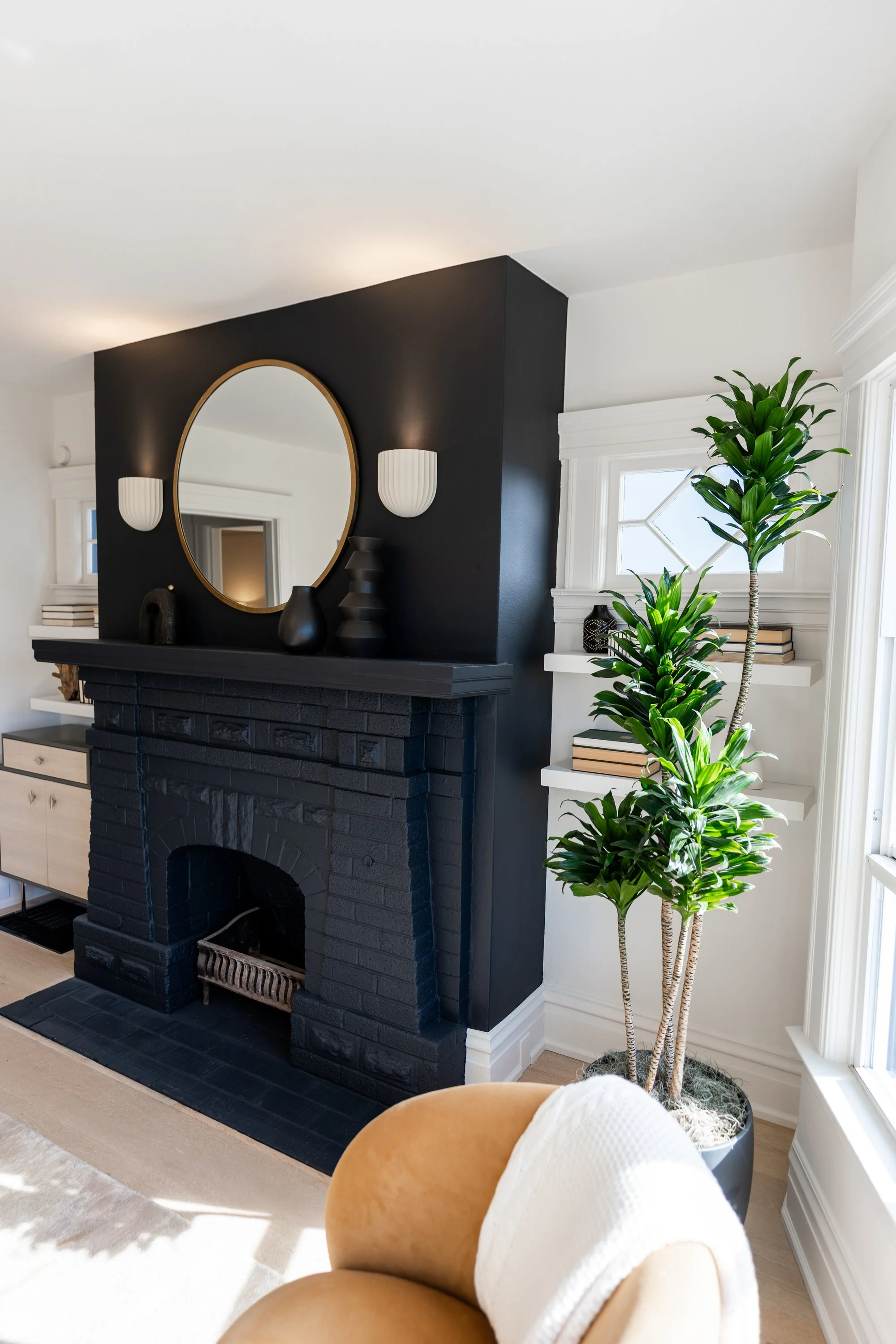 Living room with a black fireplace, a large oval mirror, white wall sconces, a tall green houseplant, built-in white shelves, decorative vases and books, and a beige armchair with a white throw blanket.