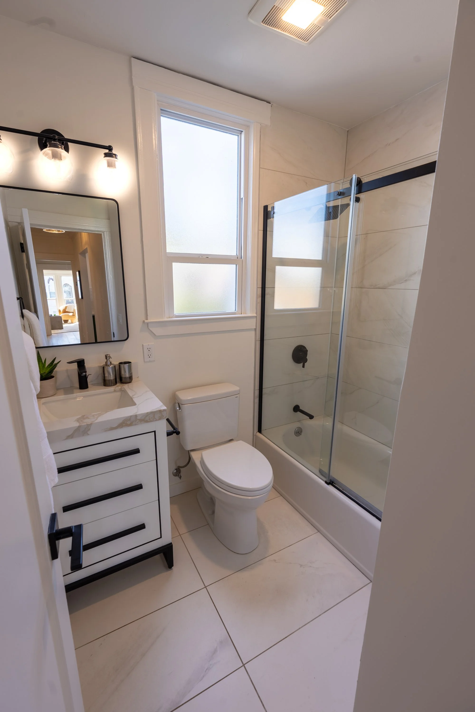 A modern bathroom featuring a white vanity with black handles, a rectangular mirror, a black faucet, a small potted plant, a soap dispenser, a toilet, a large frosted window, and a walk-in shower with beige marble tiles and a black-framed glass door.