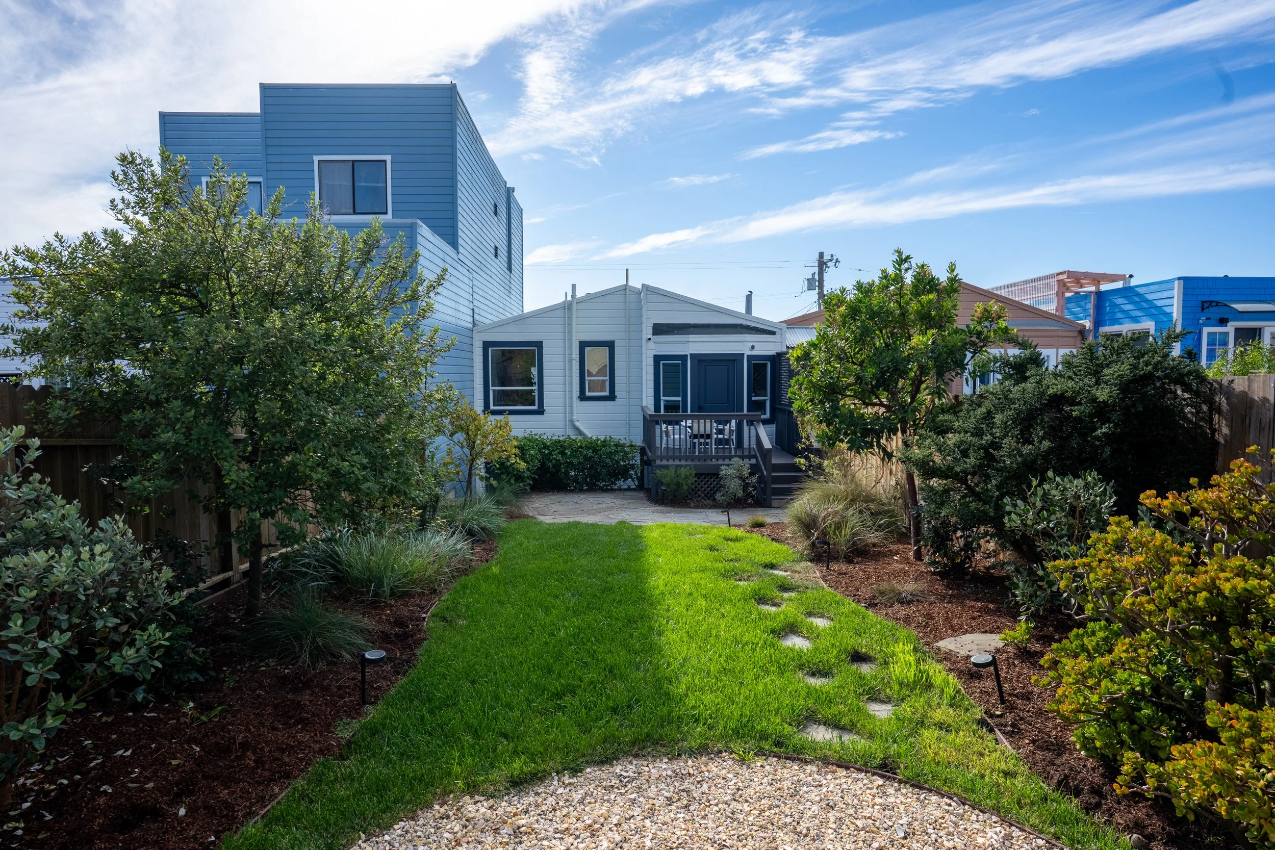 Backyard garden with lush green grass, various shrubs and trees, a stone pathway, and neighboring houses with blue and white exteriors under a partly cloudy sky.