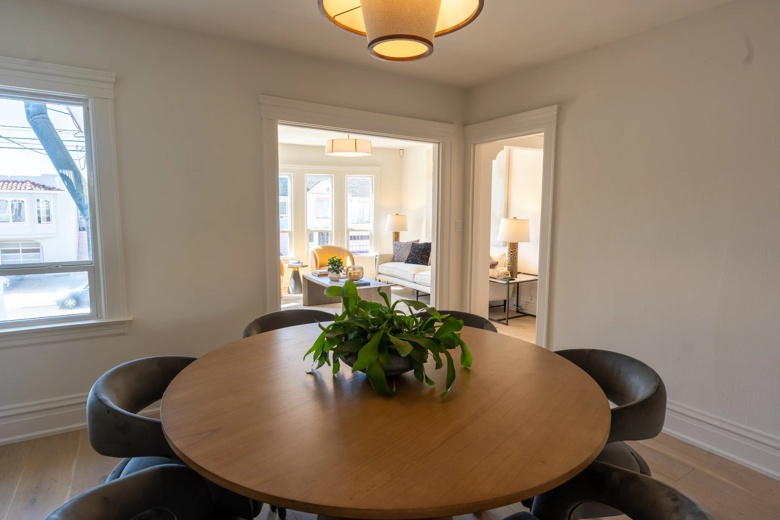 A round wooden dining table with black chairs surrounding it, a potted green plant in the center, and a view into a bright living room with white walls, large windows, and multiple lamps.