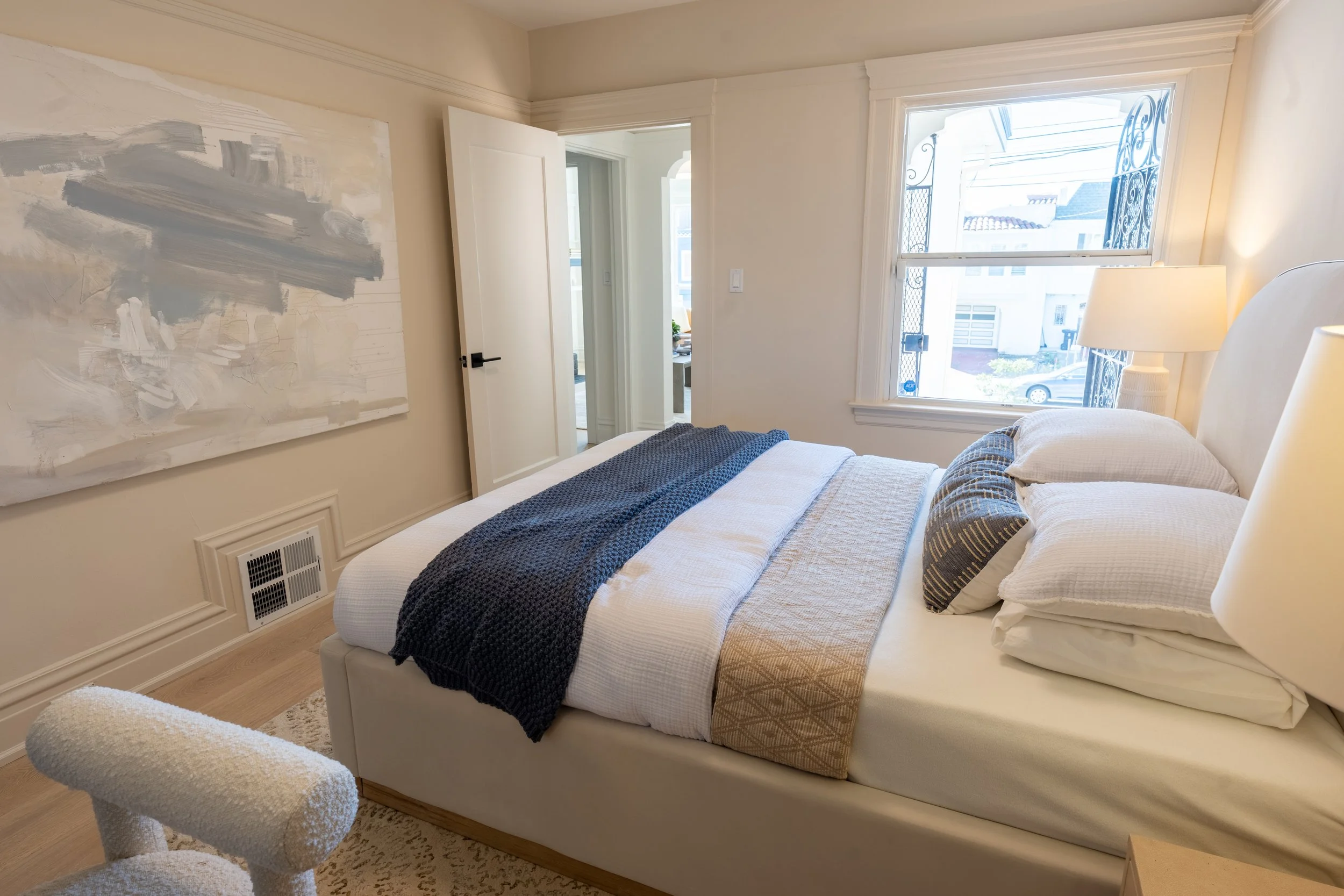 A bedroom with a neatly made bed featuring white linens, decorative pillows, and a dark blue blanket. A large abstract painting with gray and white brushstrokes hangs on the wall above a vent. There is a fluffy armchair in the foreground. The room ha