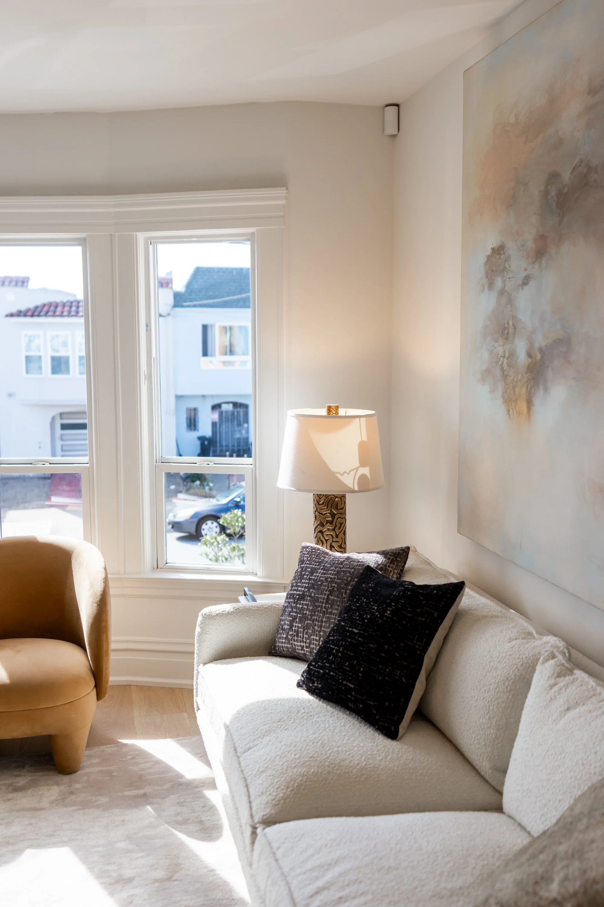 A cozy living room with a white sofa, decorative pillows, a lamp, a window with a view of neighboring houses, and an abstract painting on the wall.