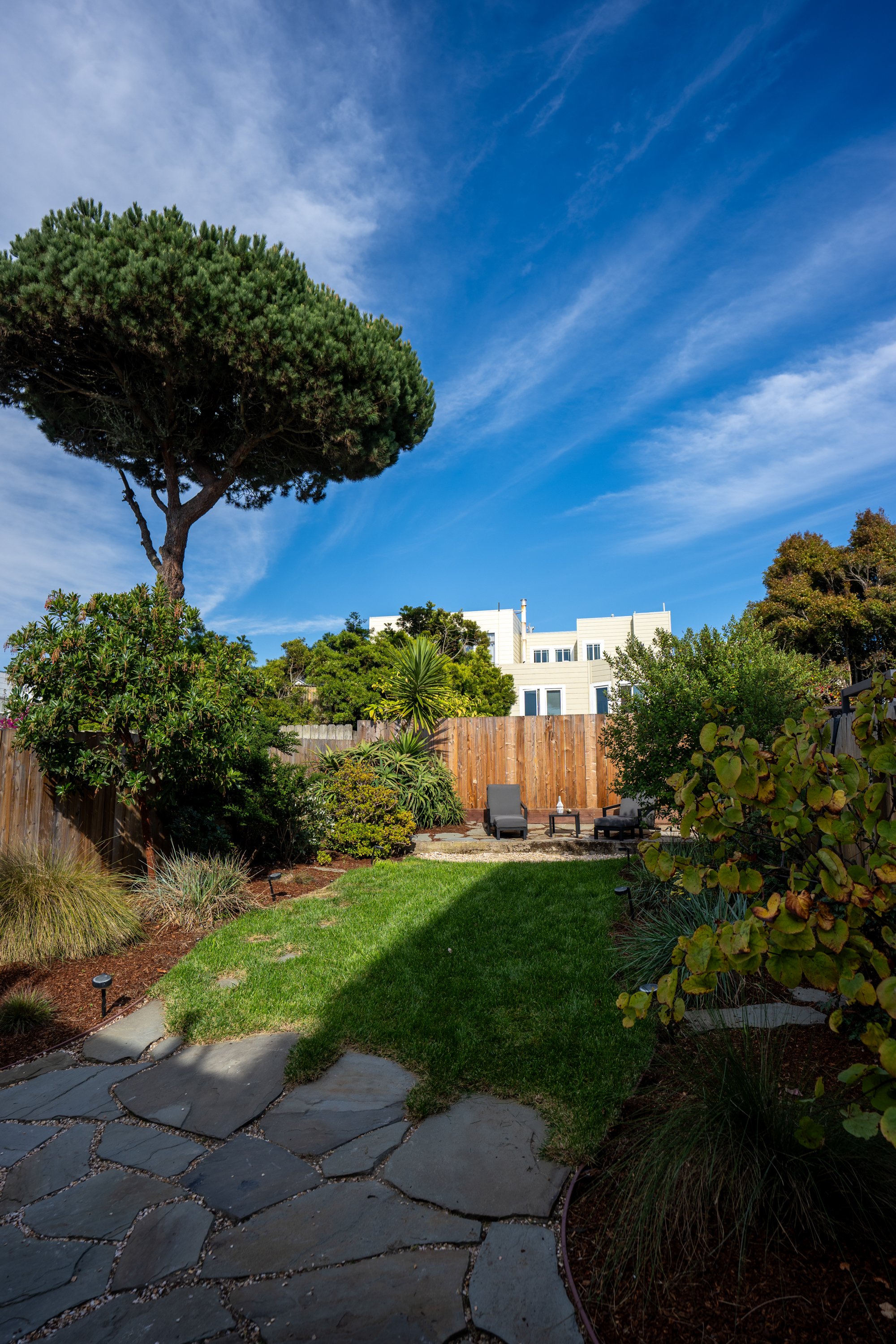 A backyard garden with a stone pathway, lush green grass, various plants and trees, and two patio chairs near a wooden fence. A large tree with dense foliage is on the left, and there are houses and a partly cloudy sky in the background.