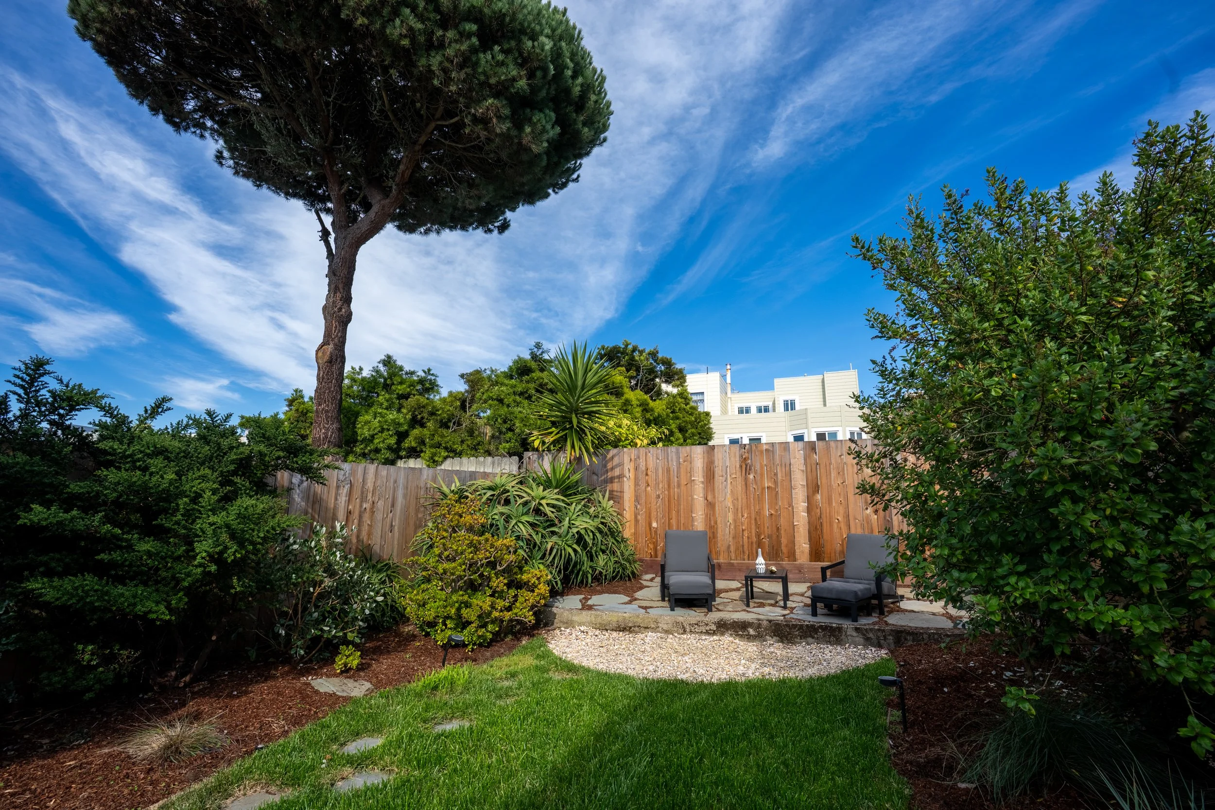 A backyard garden with lush green plants, a stone patio with two gray chairs and a small table, a wooden fence, various trees and shrubs, and a large tree with a thick trunk, against a background of a clear blue sky with wispy clouds.