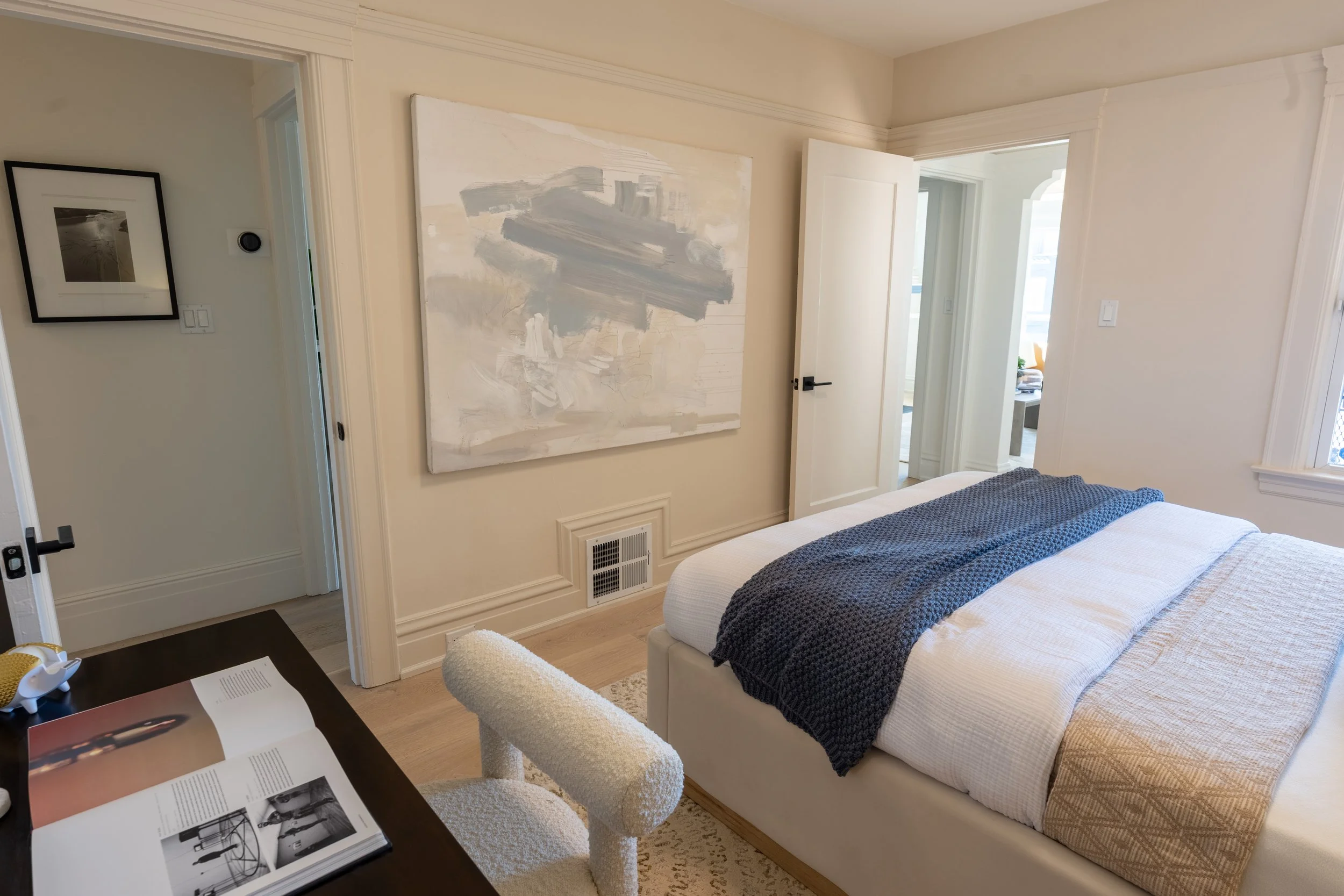 A bedroom with a white bed and a dark gray blanket, an abstract painting on the wall, a door partially open leading to another room, and a window with natural light.