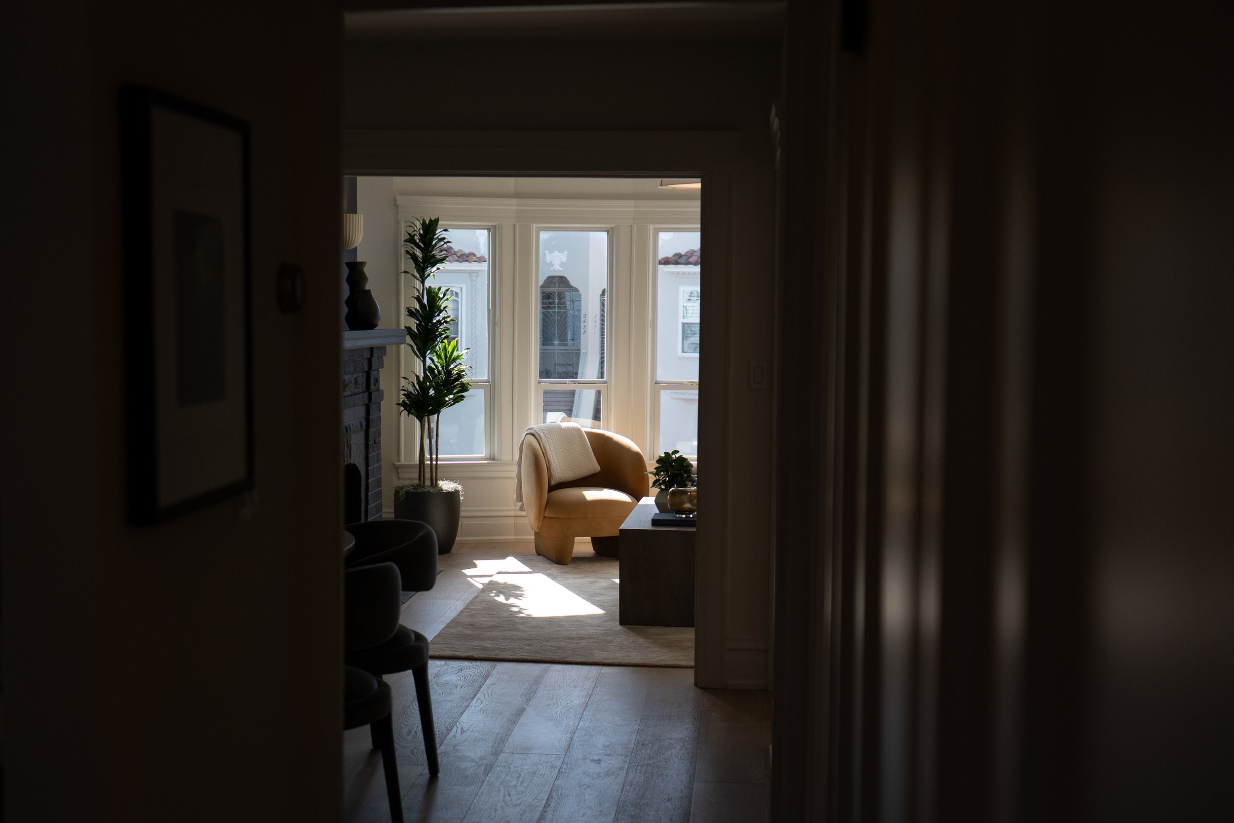 View of a cozy living room seen through a doorway, with large windows letting in natural light, a yellow armchair, a potted plant, and decorative items.