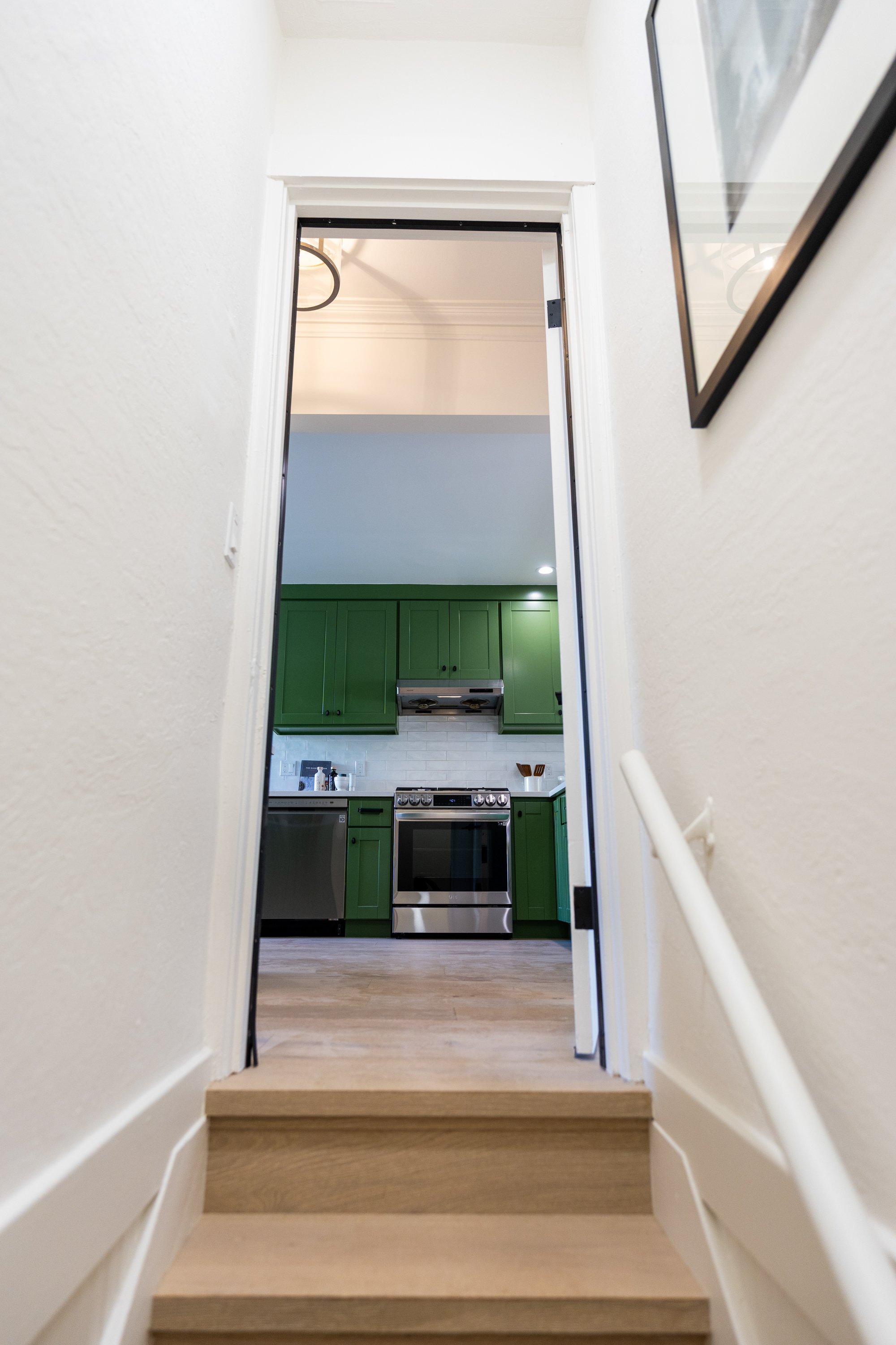 View of a kitchen area through an open doorway from stairs, featuring green cabinets, a stainless steel oven, a range hood, and a white tiled backsplash.