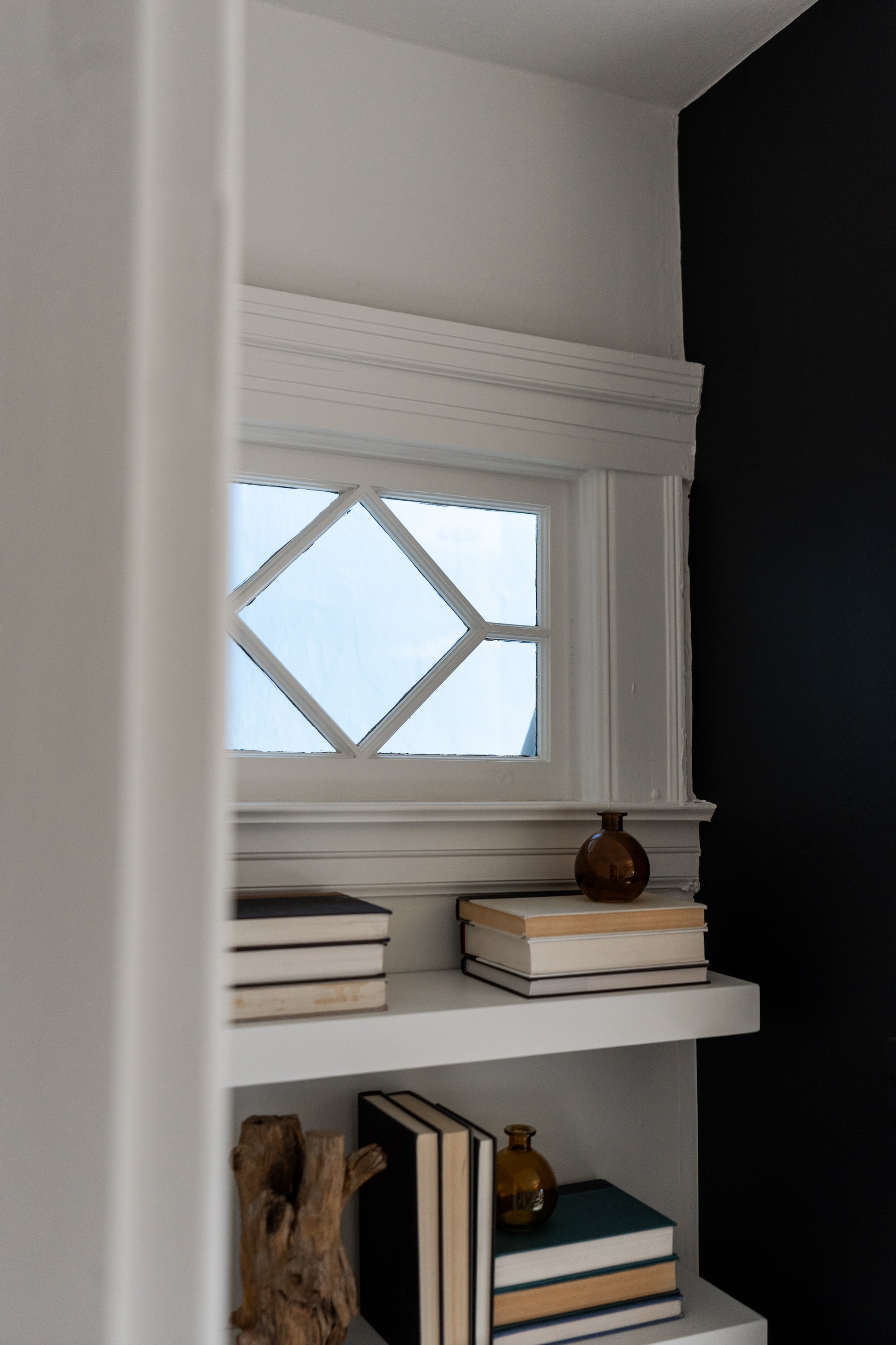 Interior view of a white bookshelf with decorative items, including books, a brown vase, and a wooden sculpture, near a window with a diamond-shaped pattern, black and white walls.