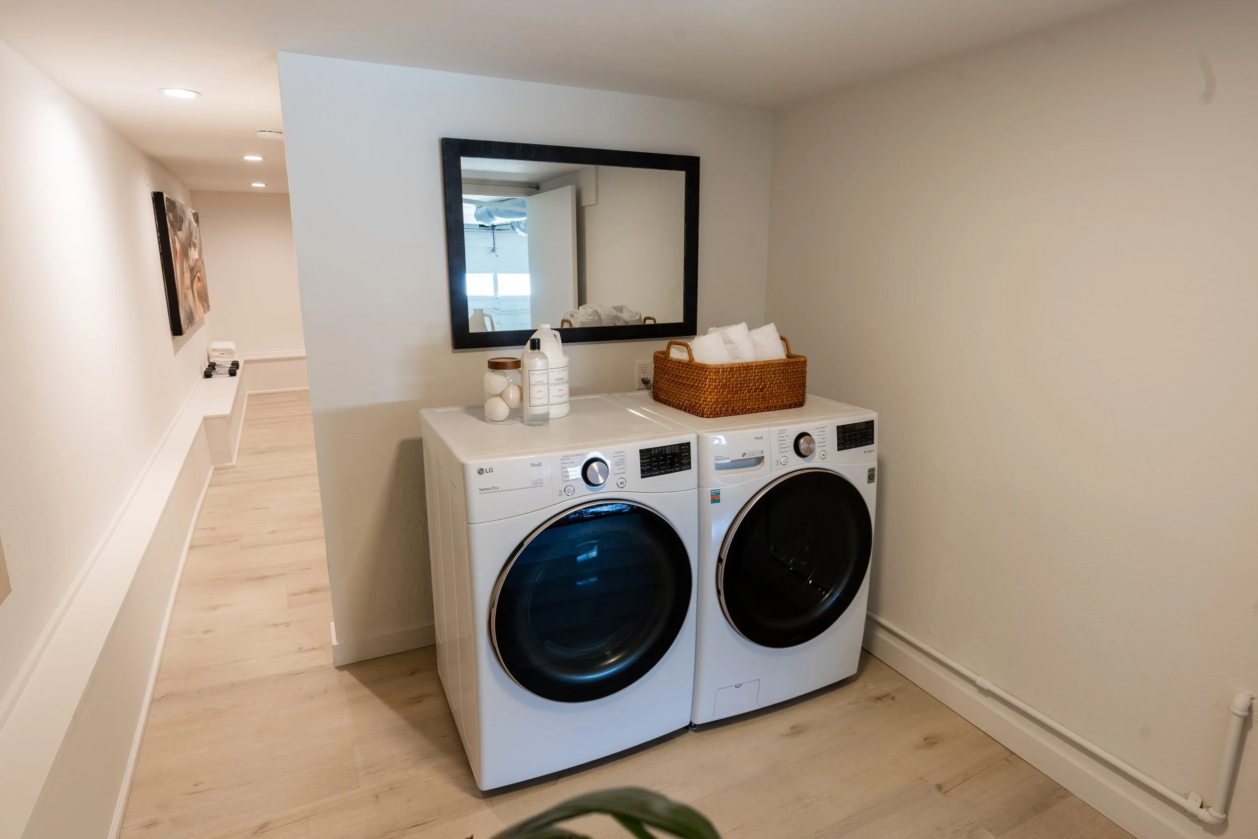 A laundry room with a white washer and dryer side by side, topped with a wicker basket containing white towels, a mirror on the wall, and some laundry supplies on top of the machines. There is a hallway with art on the wall leading out of the room.