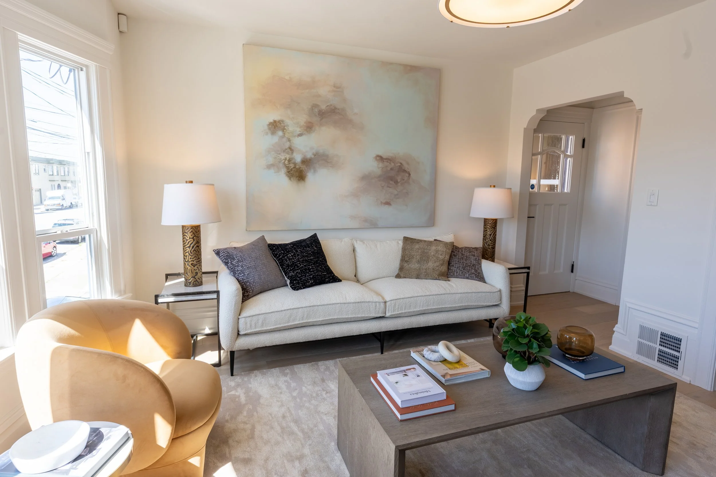 A cozy living room with a white couch decorated with black and beige pillows, a wooden coffee table with books and a plant, two table lamps, large windows, an abstract painting on the wall, and a beige armchair in the foreground.