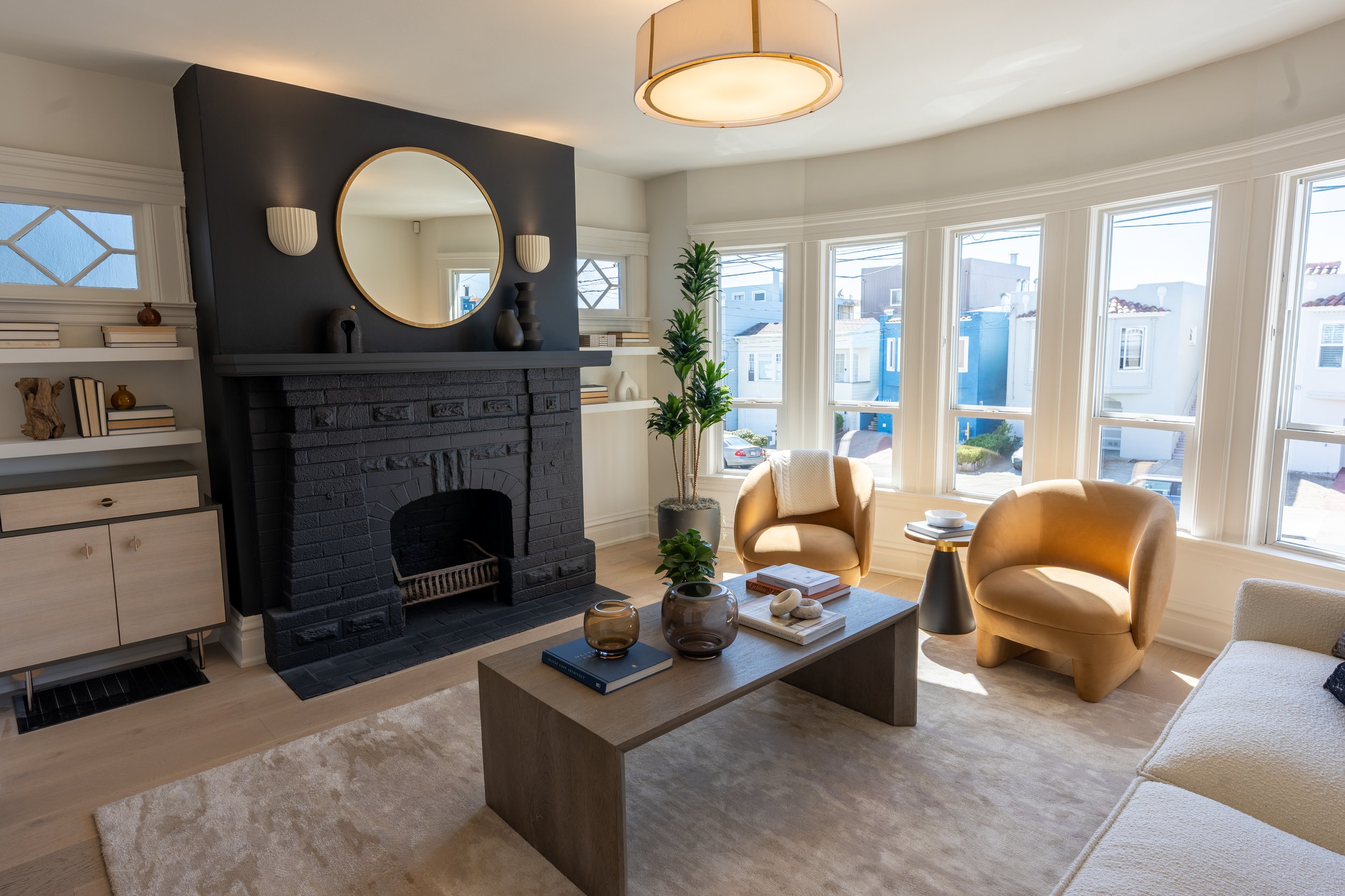 Bright living room with large bay windows, yellow armchairs, a coffee table with books and decorative objects, a black fireplace with a circular mirror above, a tall green plant, and minimalist decor.