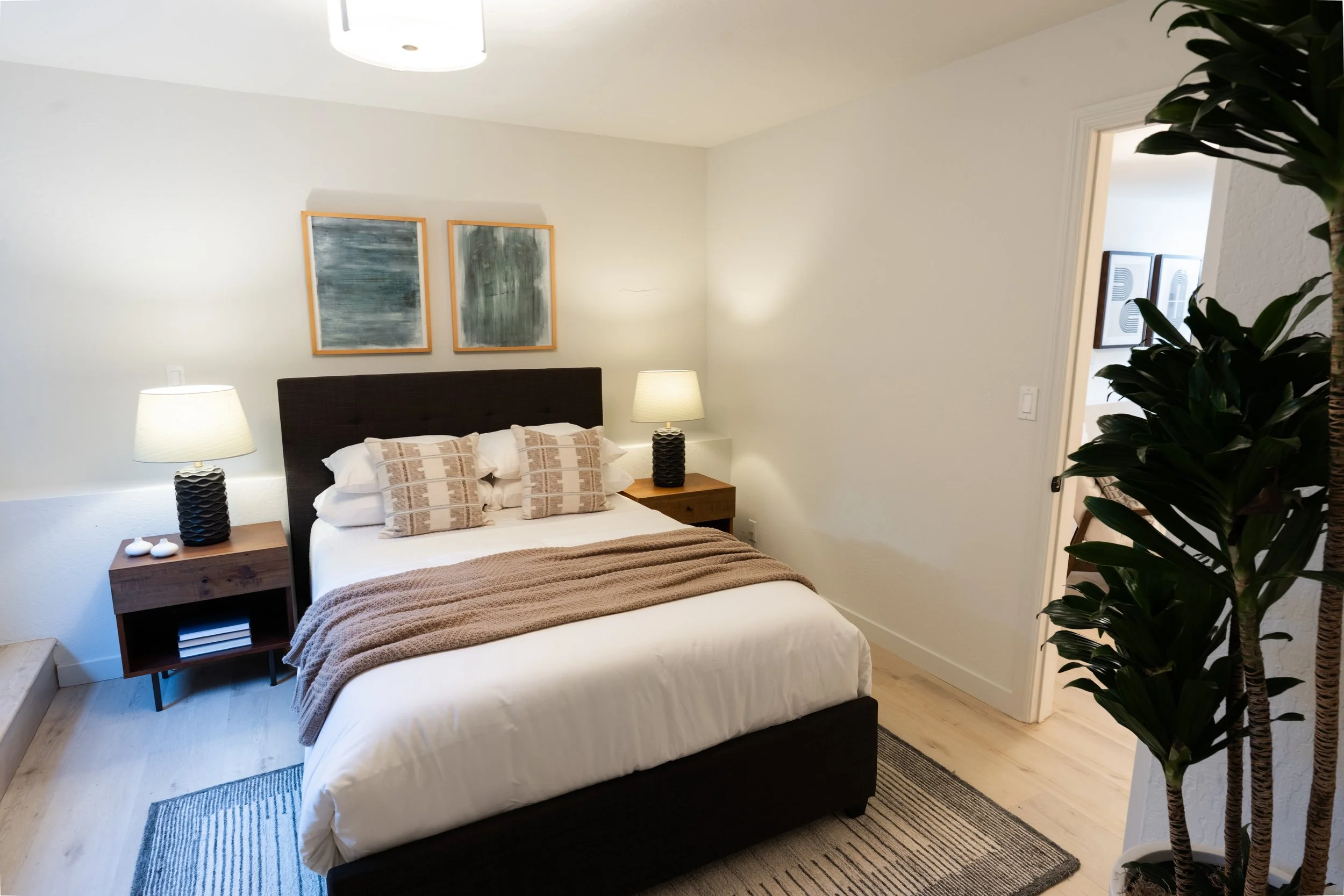 A cozy bedroom with a neatly made bed, white linens, and decorative pillows. Two nightstands with matching lamps sit on either side of the bed. Two abstract paintings hang above the headboard. A large plant is visible on the right side of the image, 