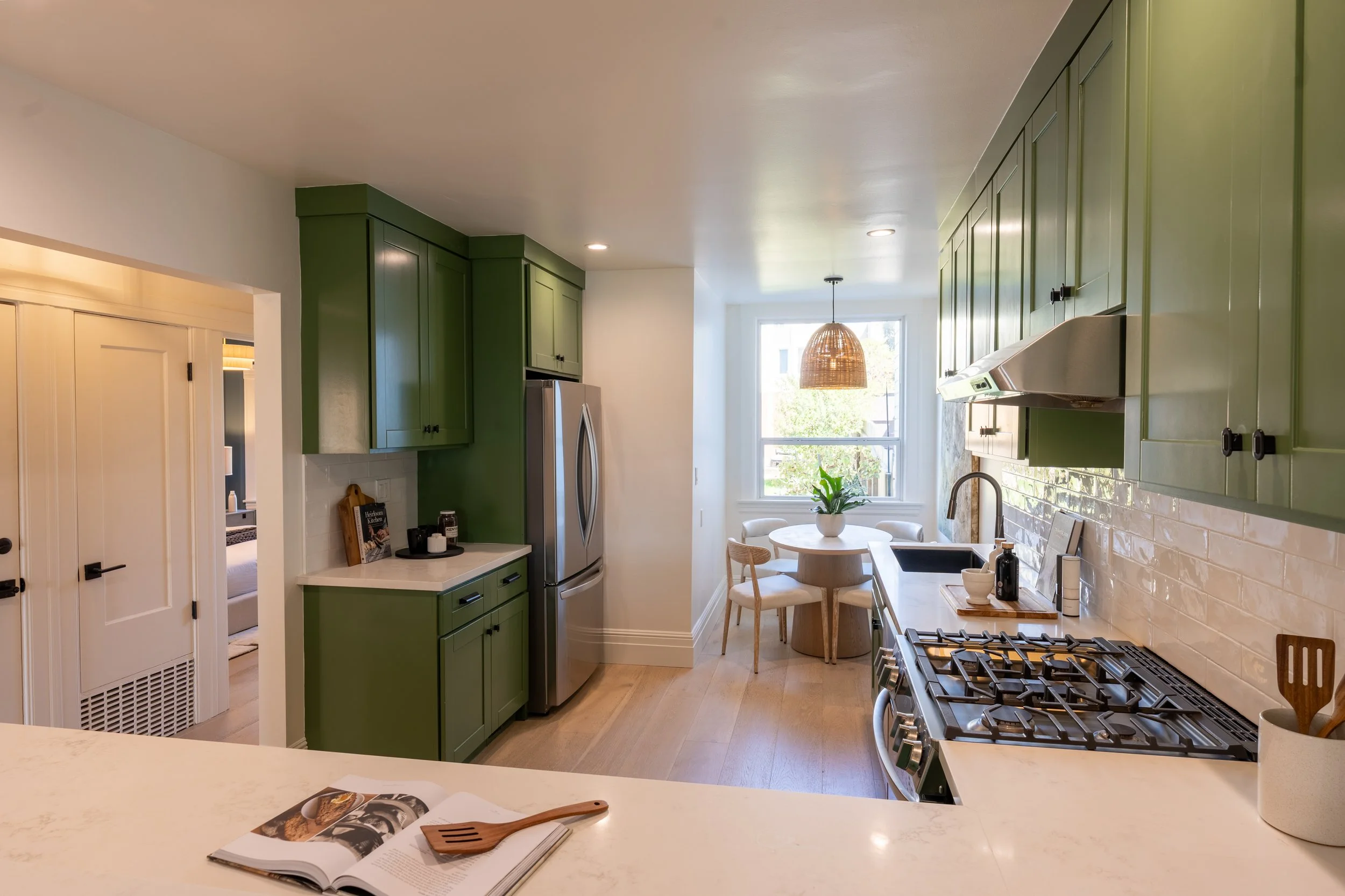 A kitchen with green cabinets, a round dining table, and a window letting in natural light. There is a stove, a refrigerator, and a plant on the table.