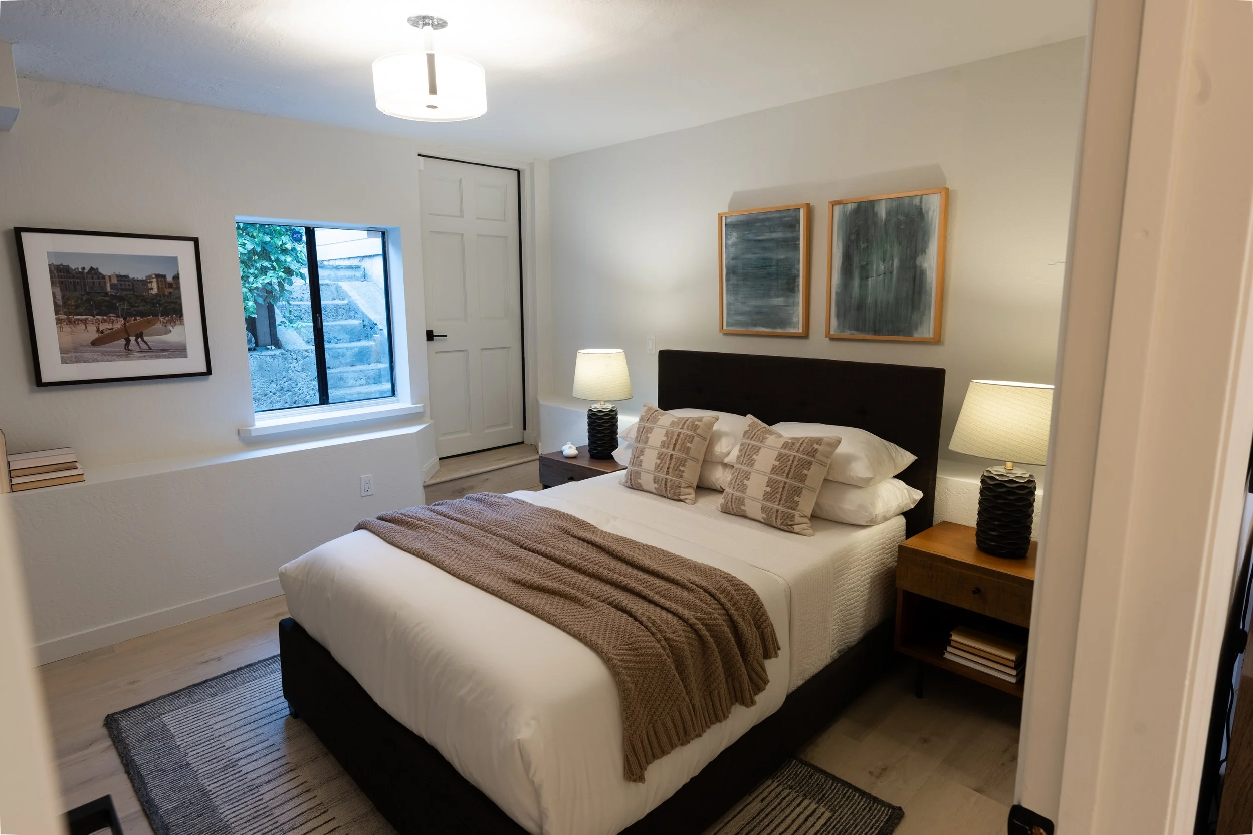 A bedroom with a bed, nightstand, and lamps, window showing steps outside, and artwork on the walls.