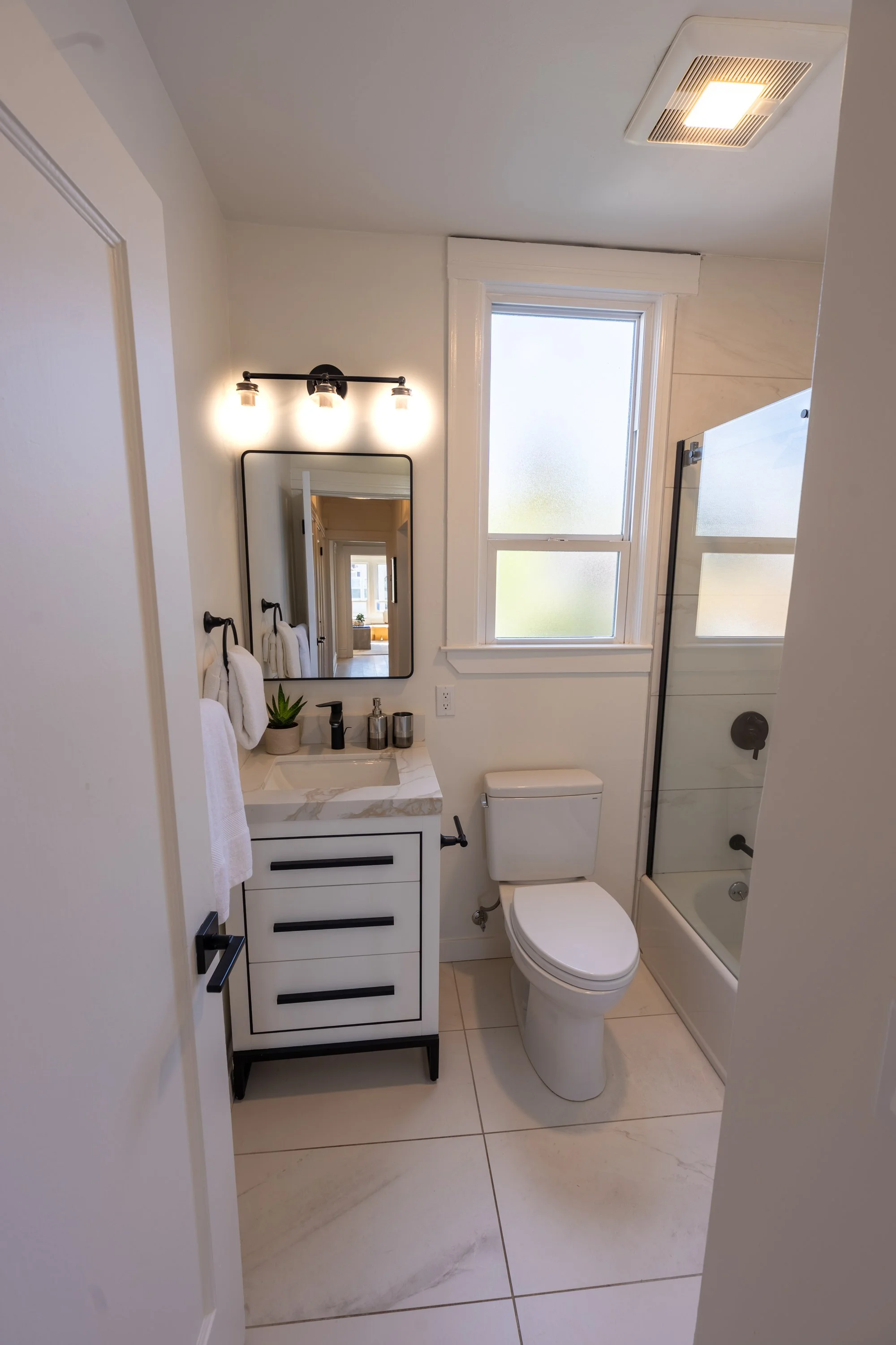 A small modern bathroom with a white vanity and marble countertop, a black-framed mirror, black fixtures, and a glass shower with a frosted window.