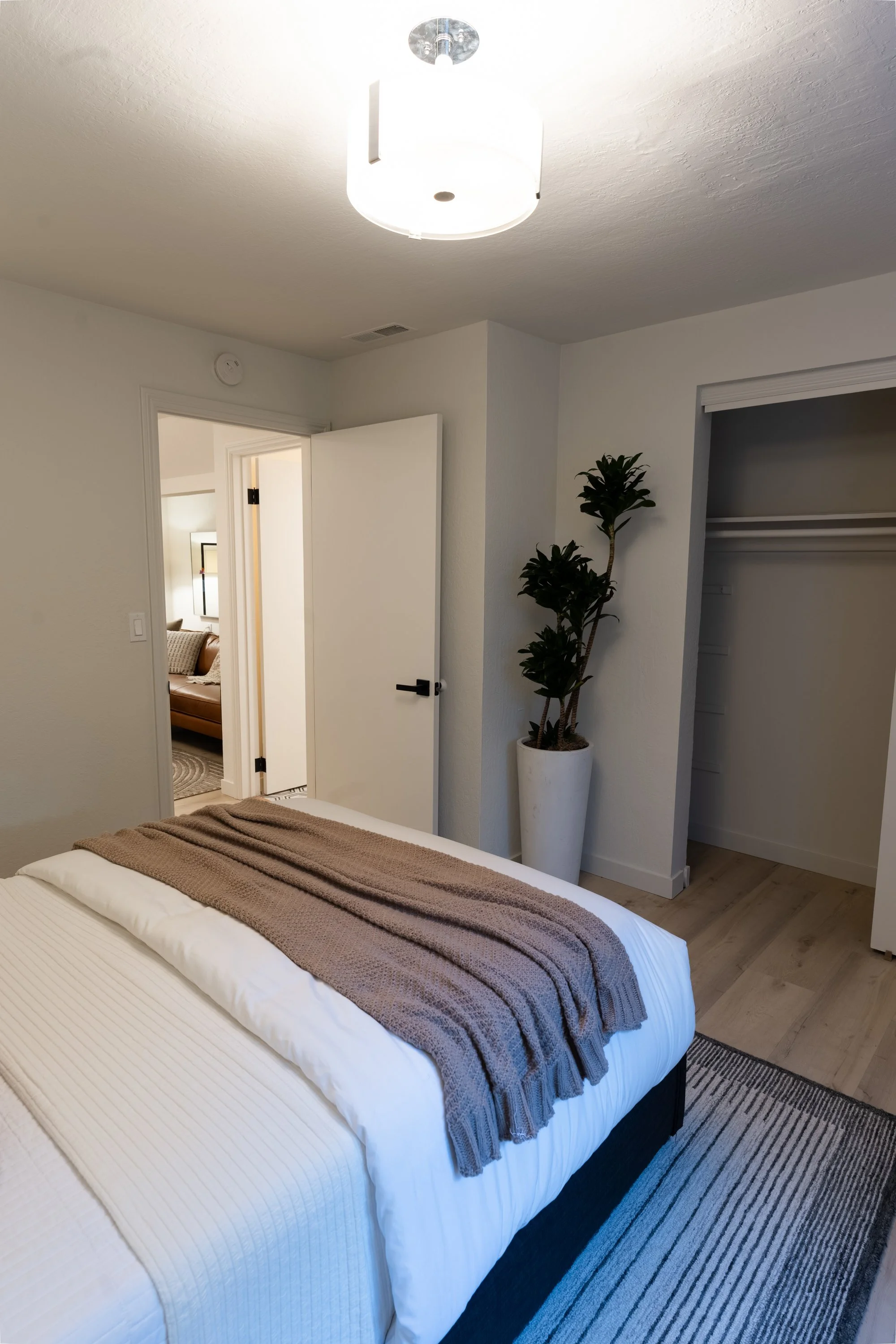 A bedroom with a bed covered in white and brown linens, a nightstand, a potted plant, and an open closet door. A ceiling light fixture is also visible.