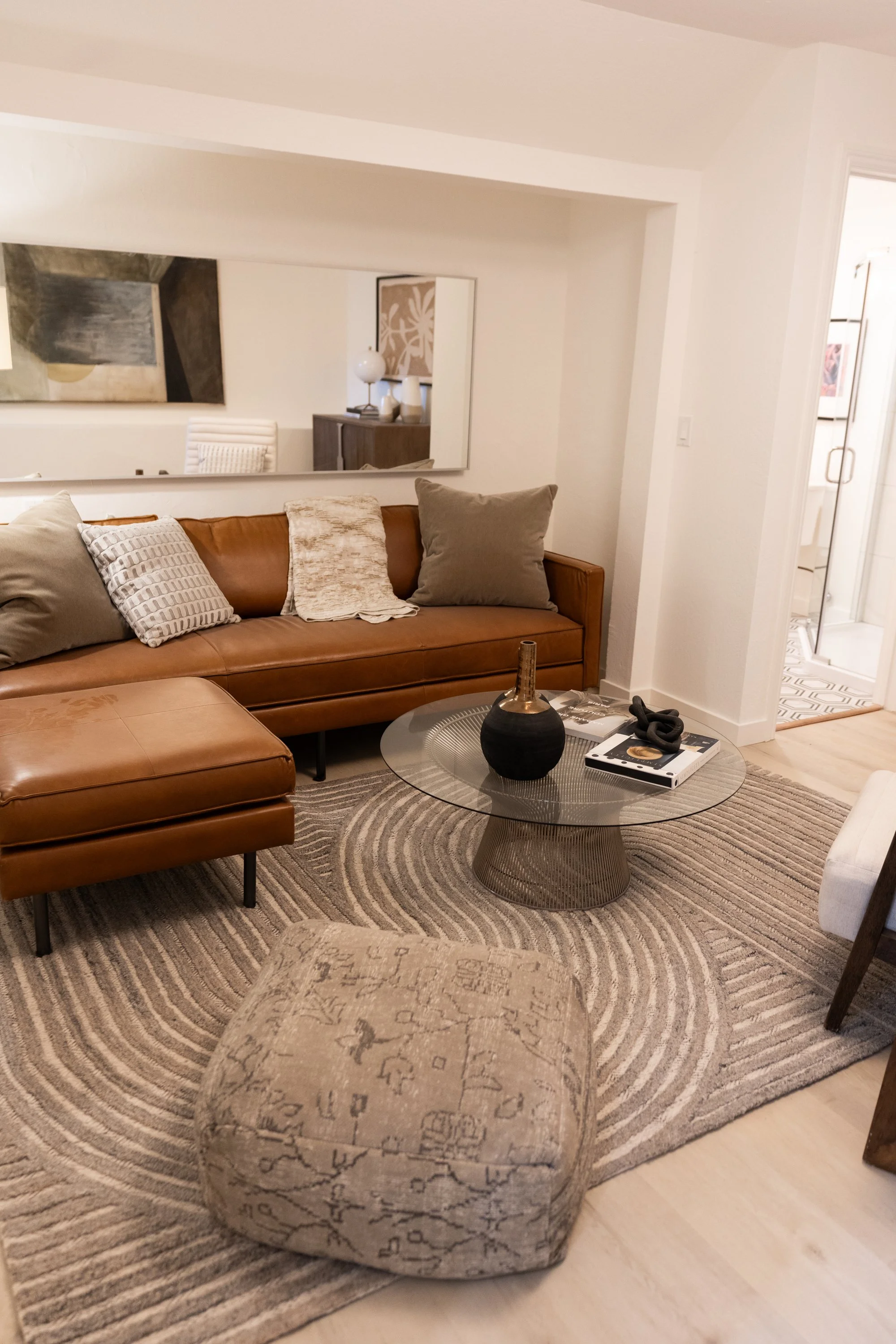 Living room with tan leather sofa, beige patterned ottoman, glass coffee table with black vase, spiral rug, and wall mirror reflecting artwork and decorative objects.