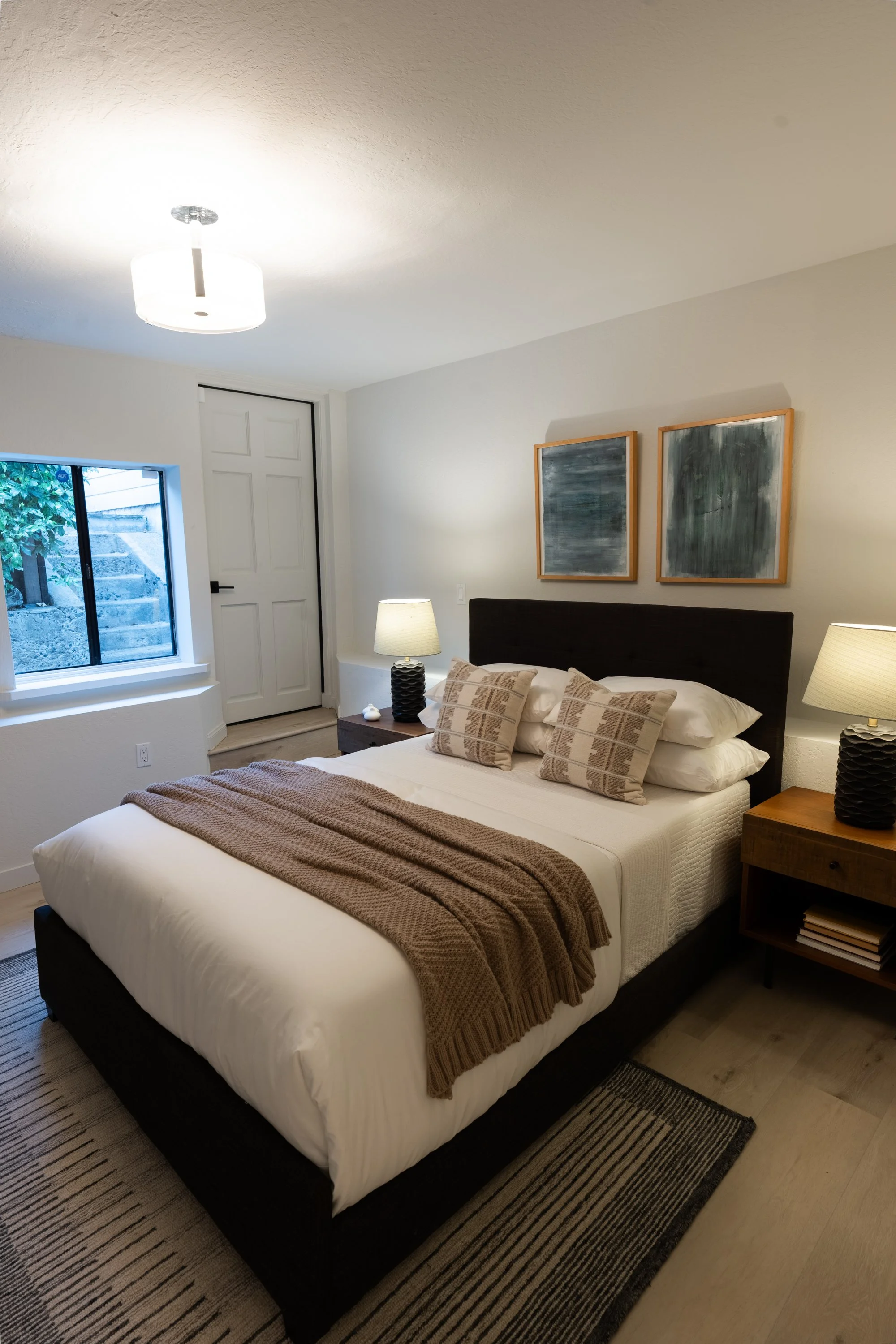 A bedroom with a bed covered in white bedding, brown blanket, and beige pillows. Beside the bed are two matching lamps on wooden nightstands. On the wall above the bed are two framed abstract art pieces. There is a window with a view of stone steps a