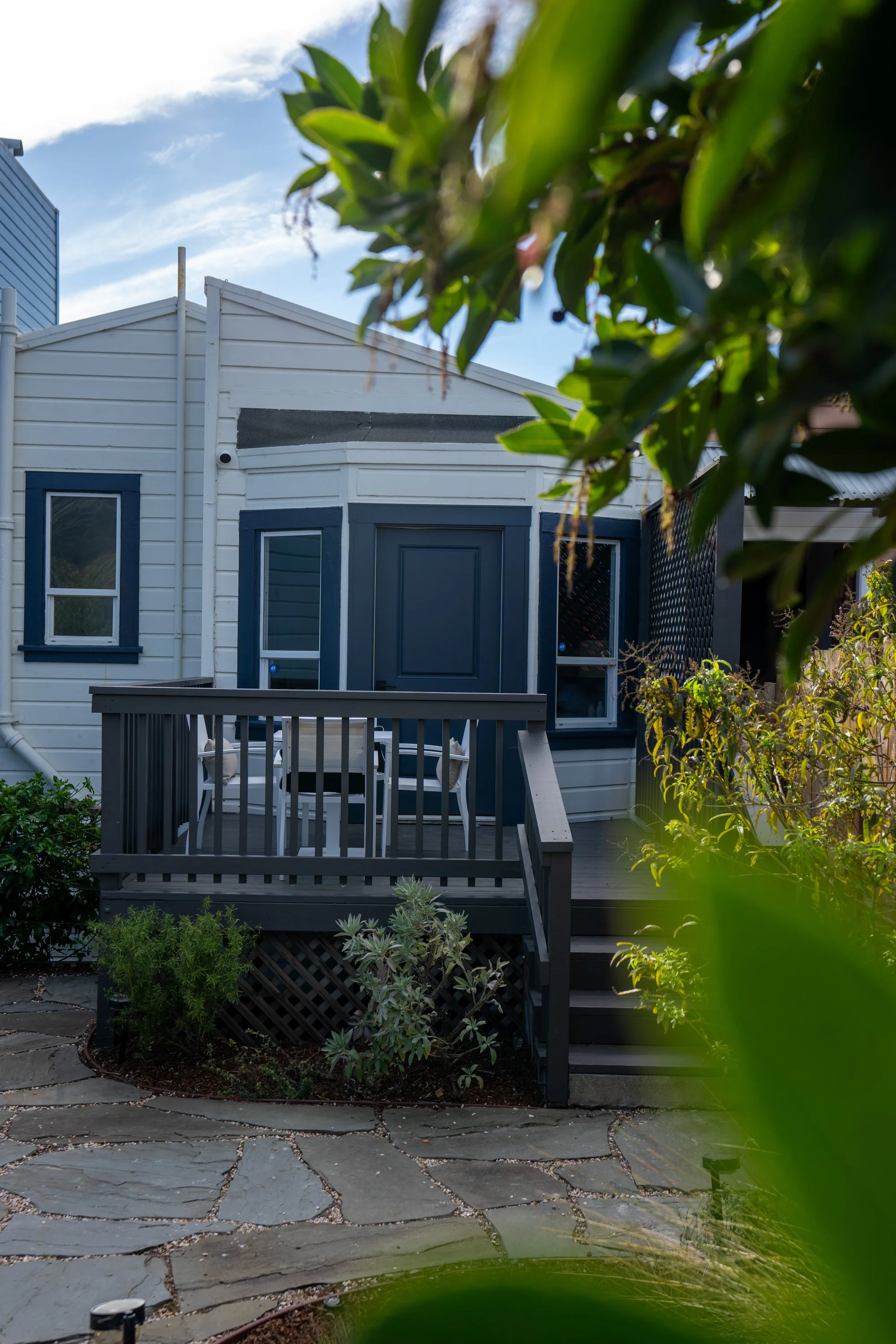 Exterior view of a small house with a dark gray porch, white siding, and blue window frames and door, surrounded by green plants and trees, with a stone pathway in front.