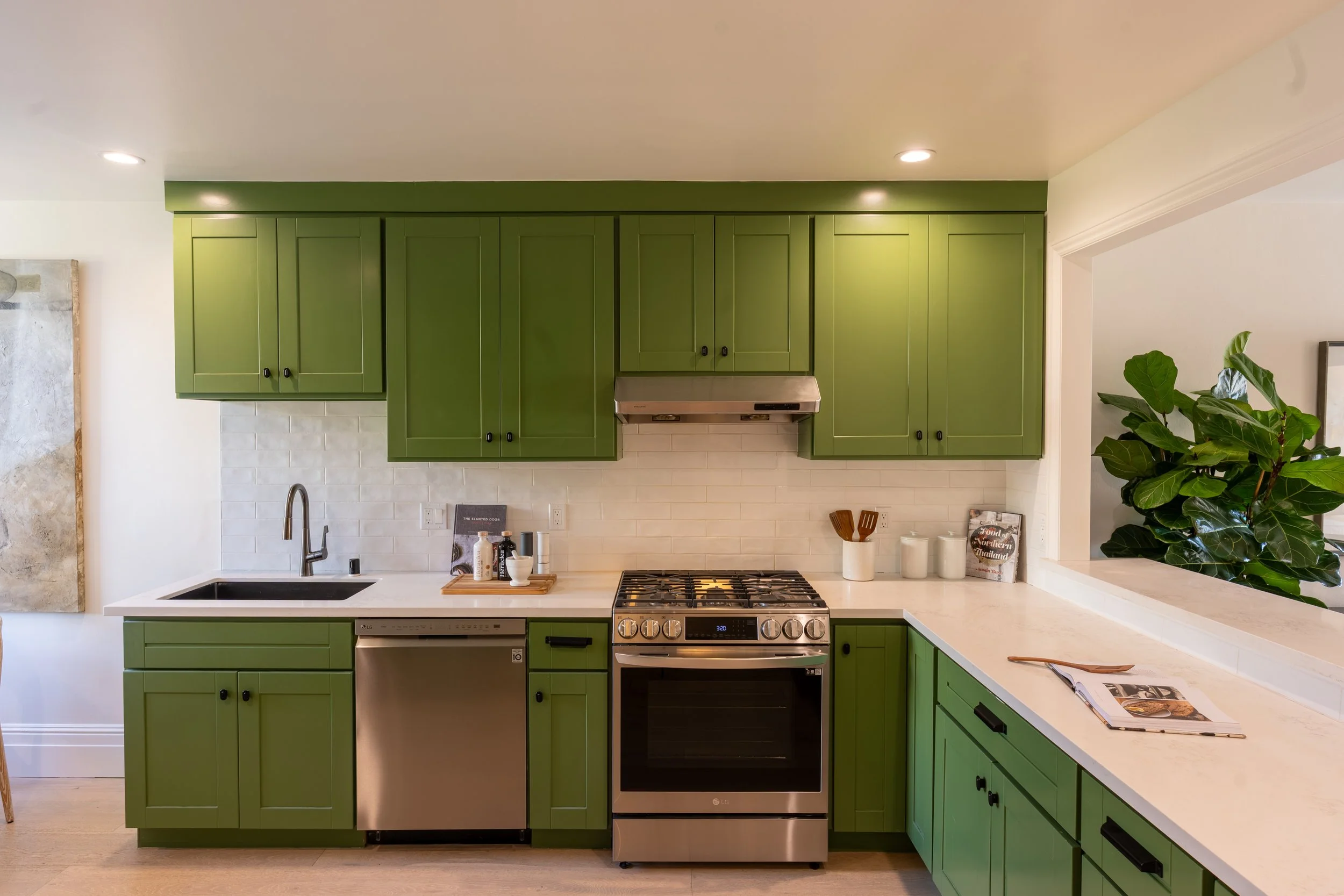 Kitchen with vibrant green cabinets, white subway tile backsplash, stainless steel oven and range, black hardware, and a white countertop with a plant and some reading material.