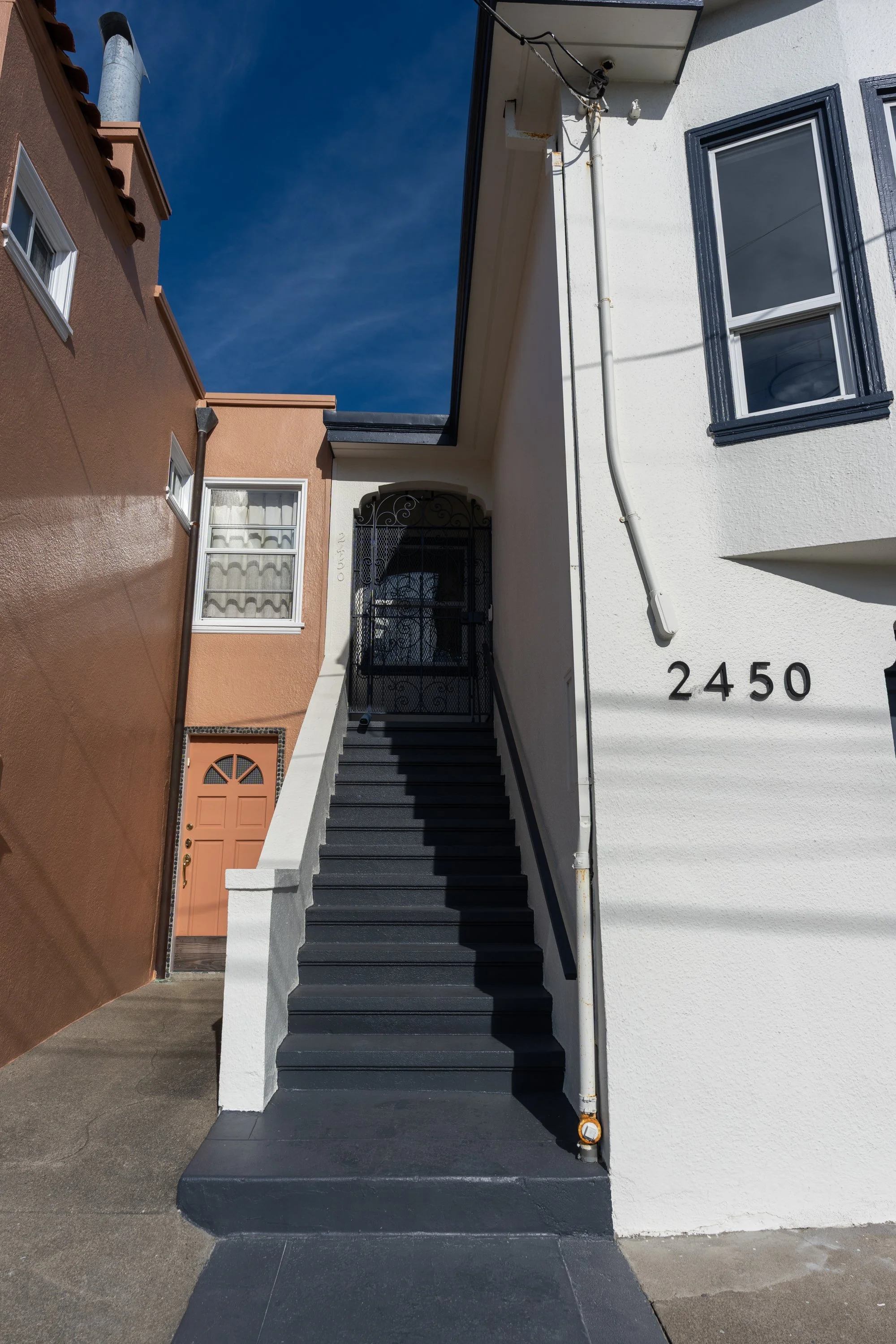 Exterior of a white house with black stairs leading to an upper door, house number 2450, and adjacent brown house with a small window. Clear blue sky in background.