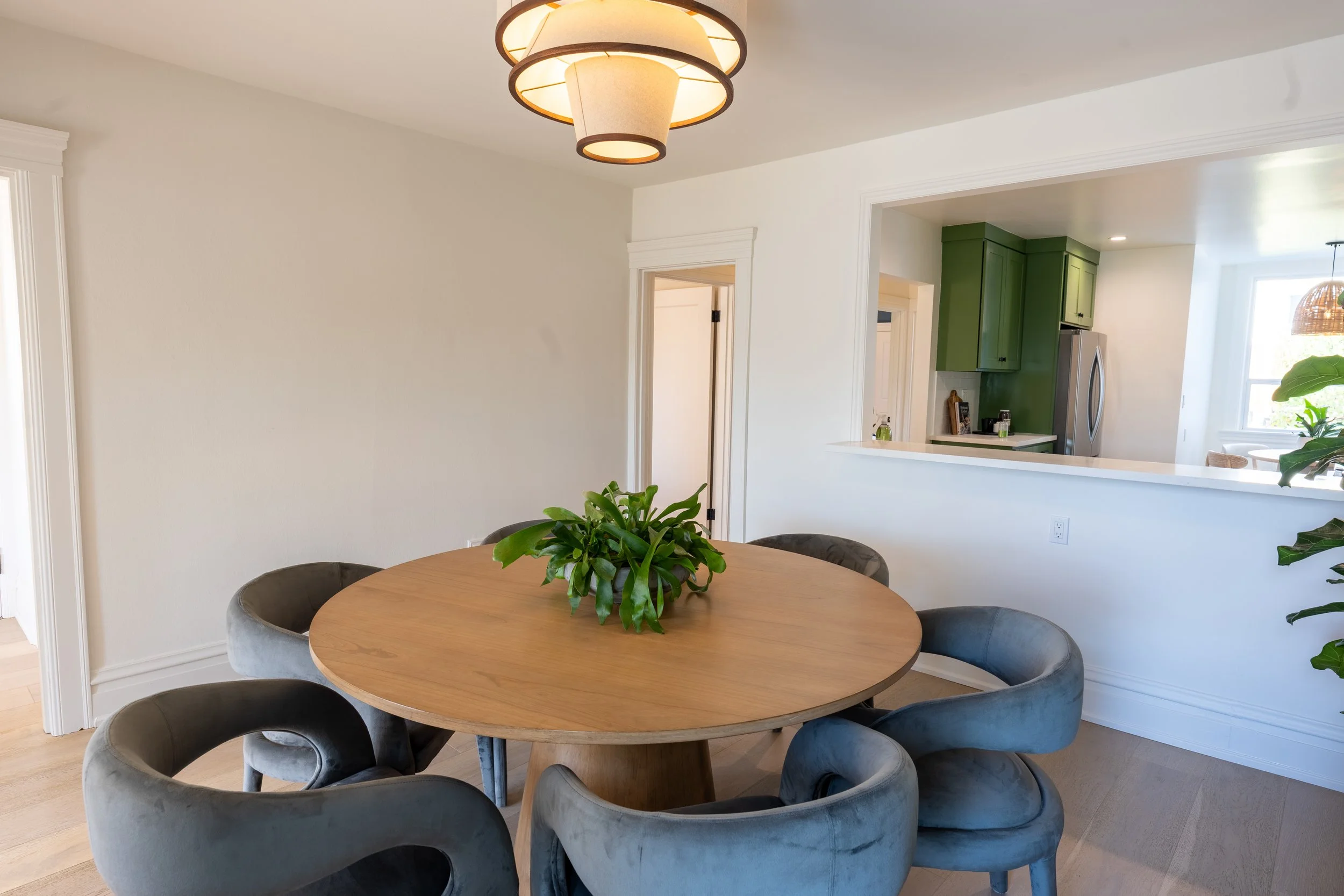 A dining area with a round wooden table surrounded by six velvet chairs. There is a potted plant on the table. In the background, a view of the kitchen with green cabinets and a kitchen counter can be seen, along with a window with a view outside.