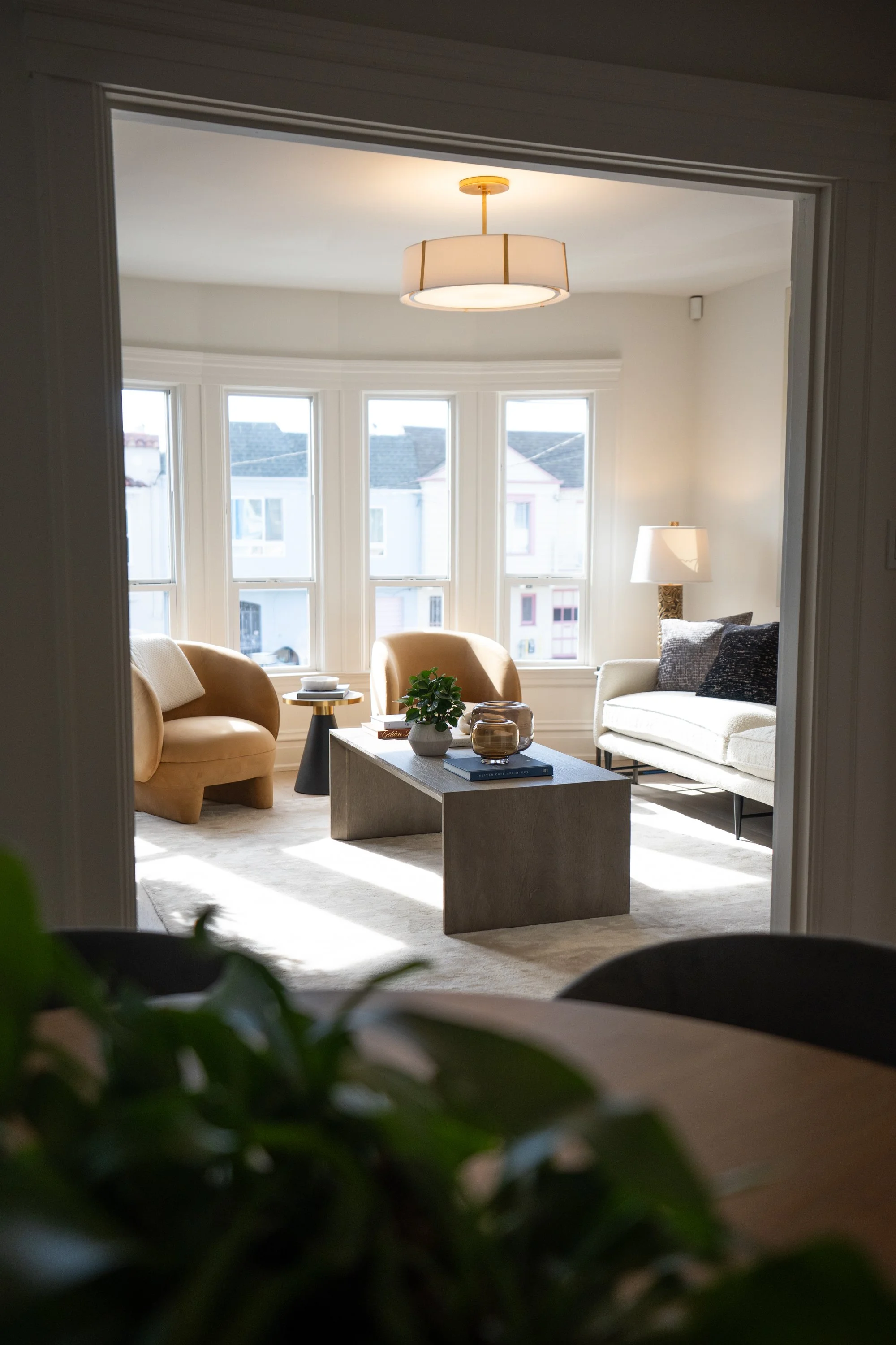 Bright living room with large bay windows, beige armchairs, white sofa, and modern lighting, viewed through an archway.