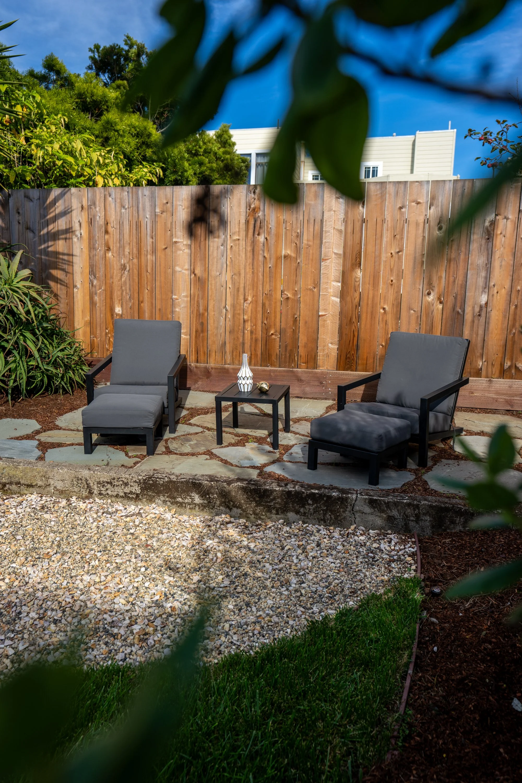 Backyard patio with two gray chairs, a small black table with decorative objects, a wooden fence, green foliage, and a blue sky.