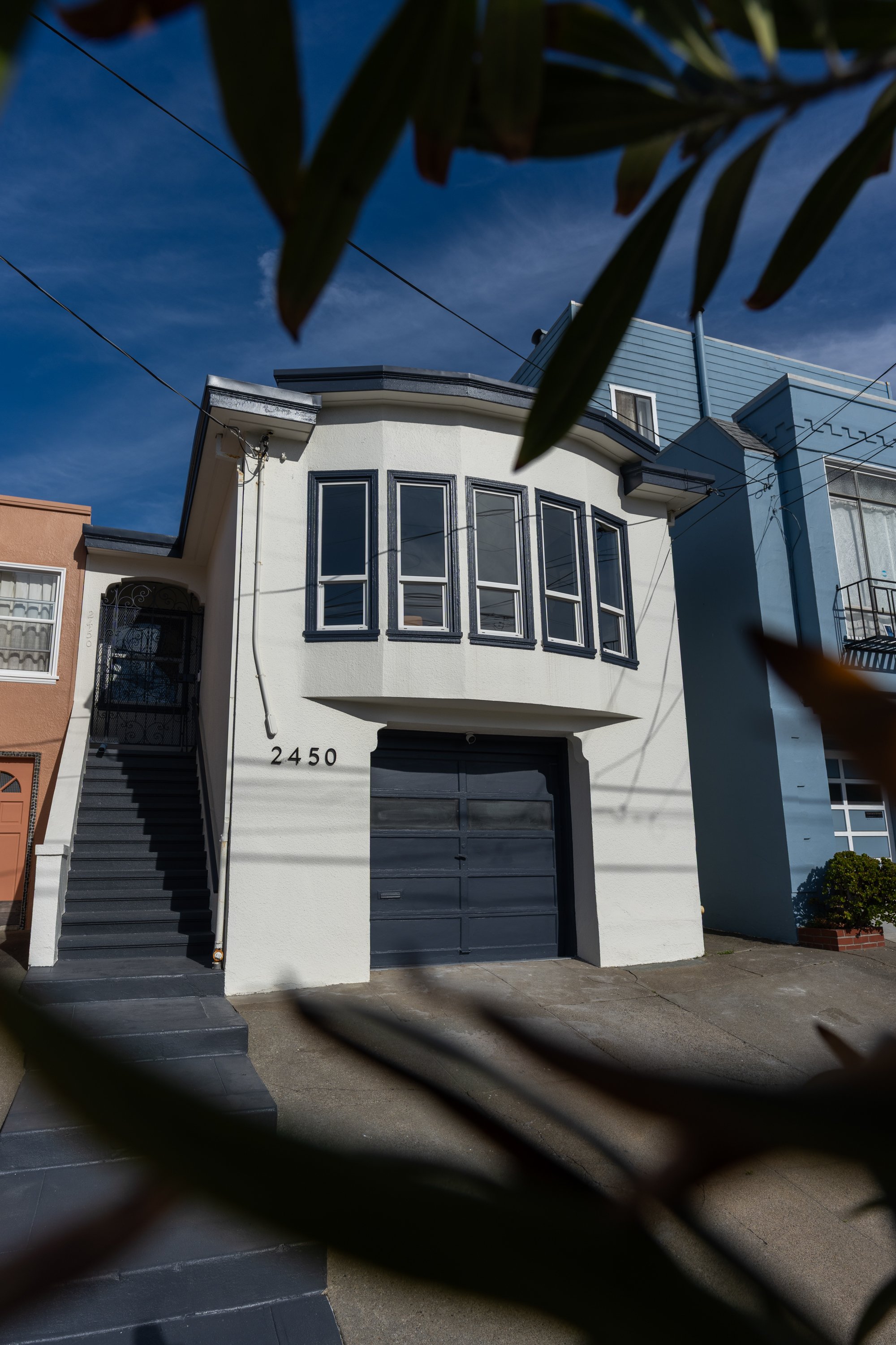 Front view of a white two-story house with black window frames and a garage door, with a staircase leading to the entrance and visible neighboring buildings, all partially obscured by leaves in the foreground.