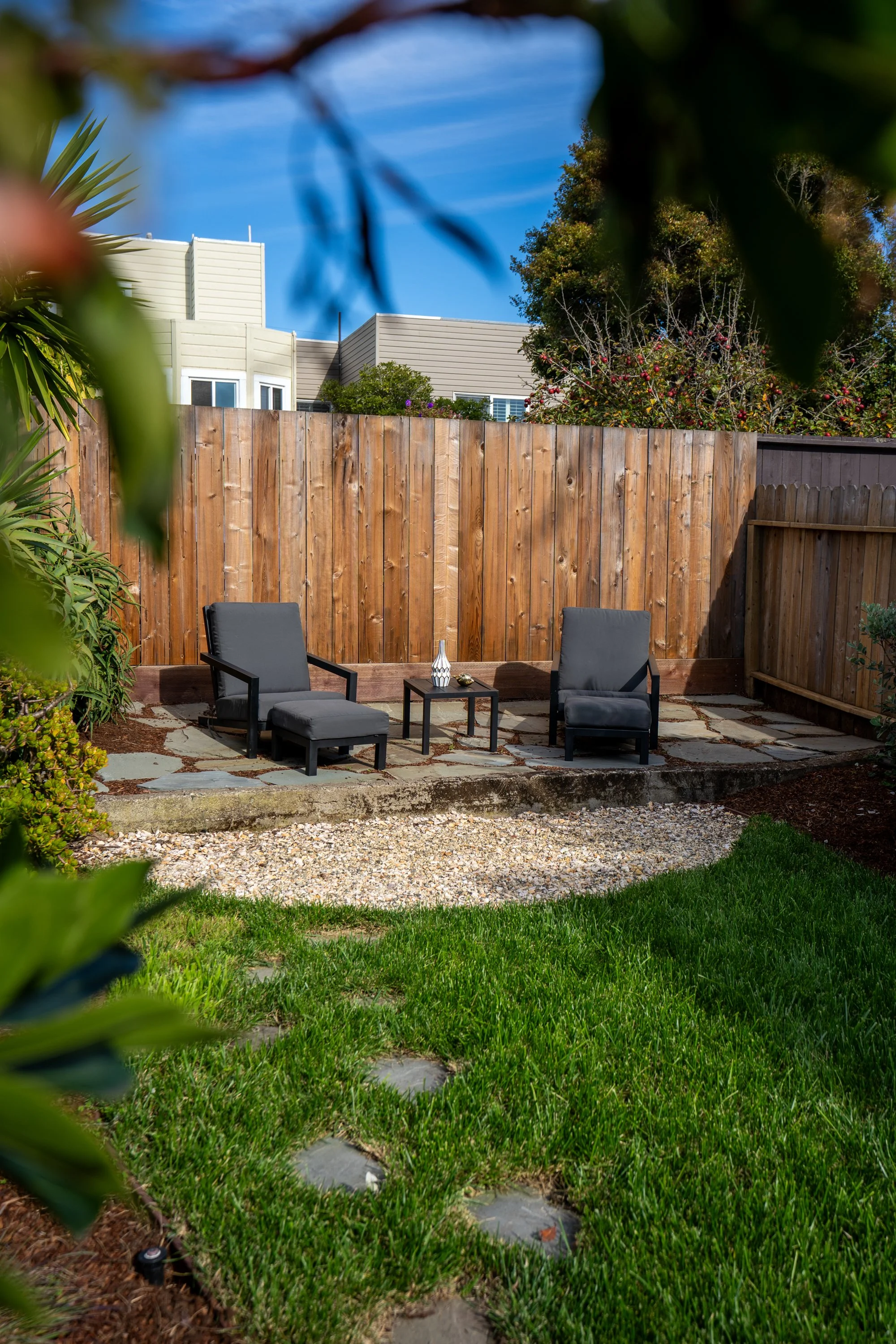 A backyard patio area with a wooden privacy fence, two black patio chairs with matching ottomans, a small table with decor, green plants and trees, a gravel pathway, and modern beige houses in the background under a bright blue sky.